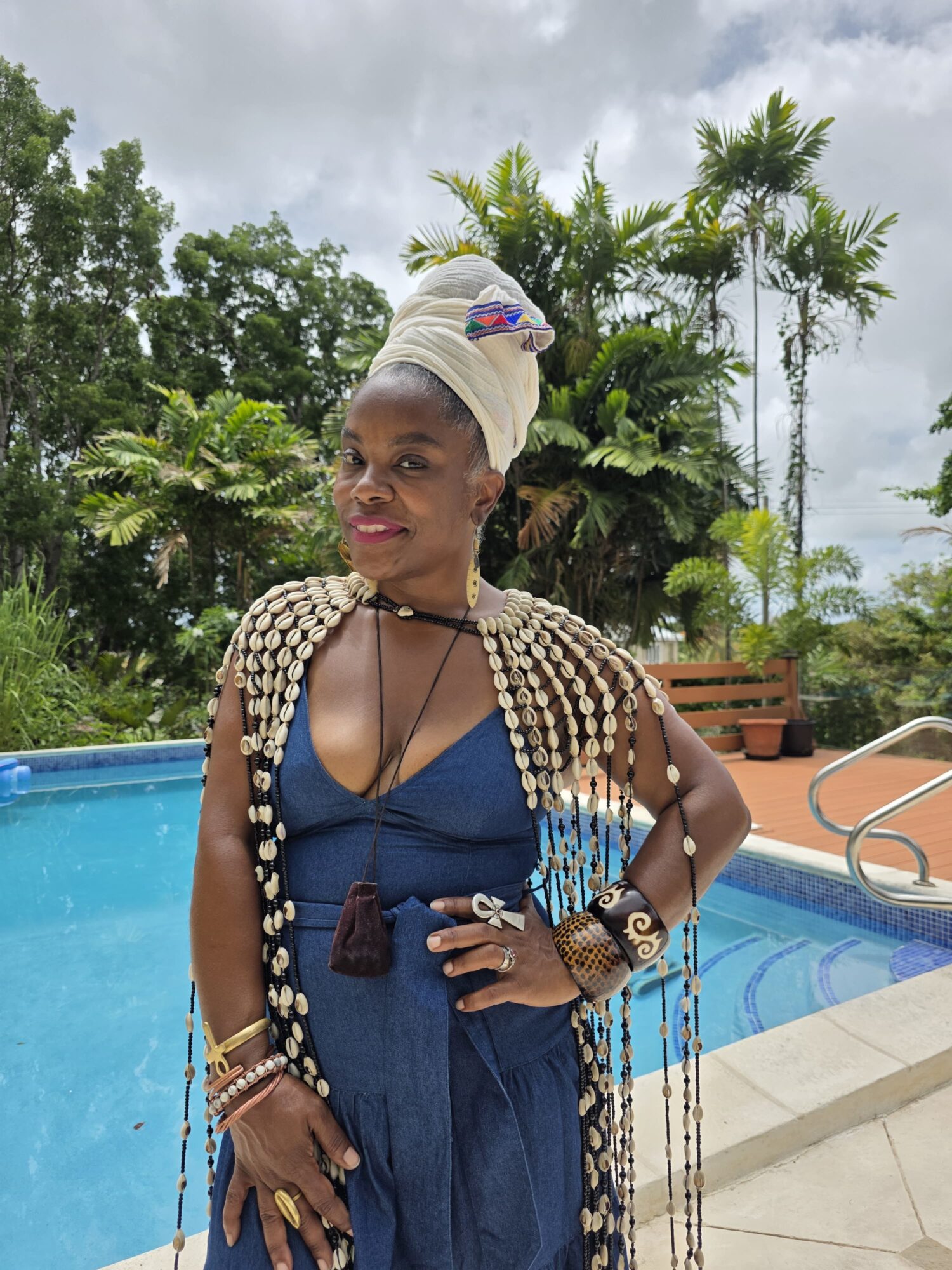 Woman in blue dress with beaded accessories and headwrap, standing by a pool with tropical trees in background.
