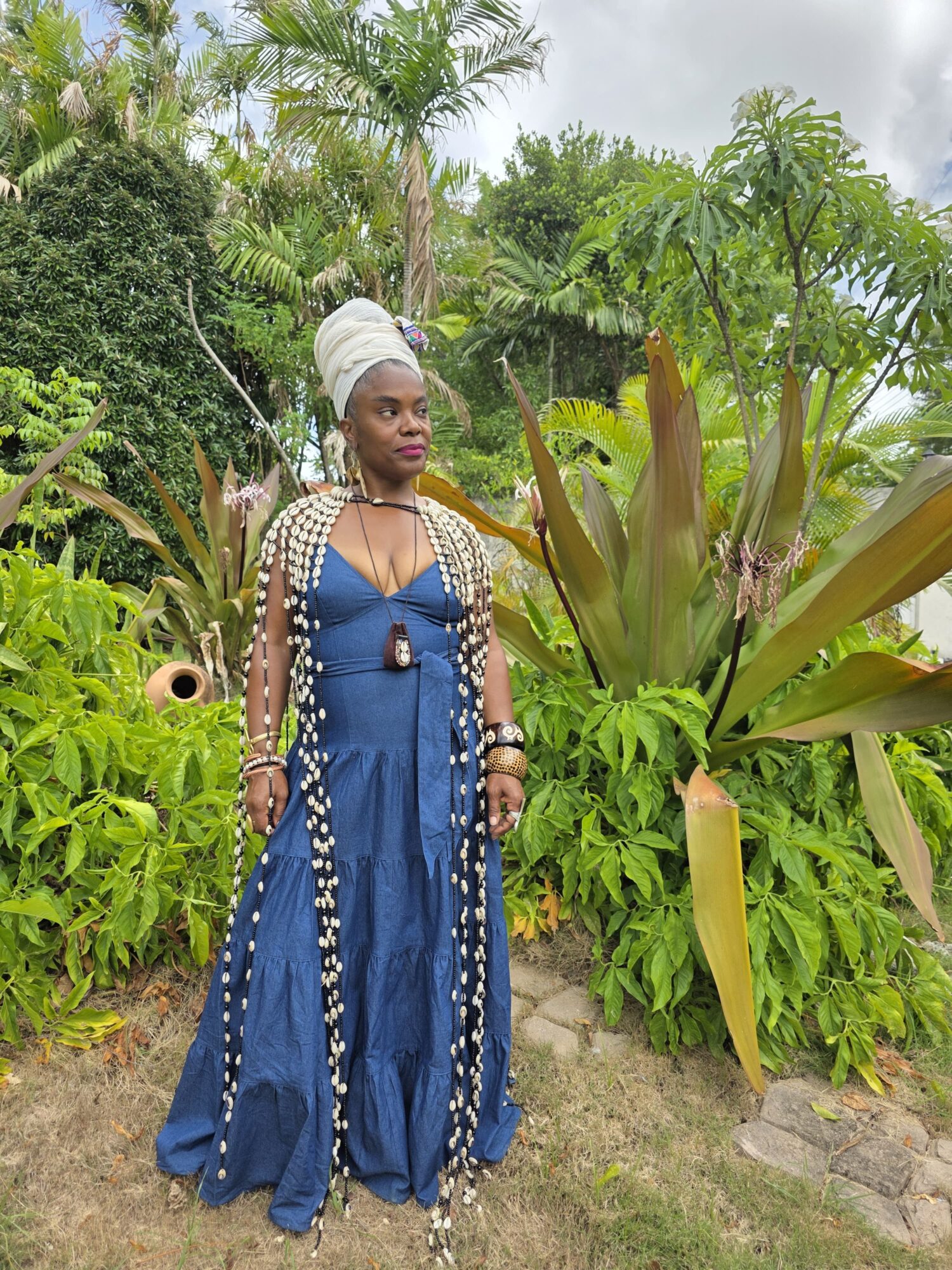 Woman in blue dress and white headwrap standing outdoors among green plants and trees.