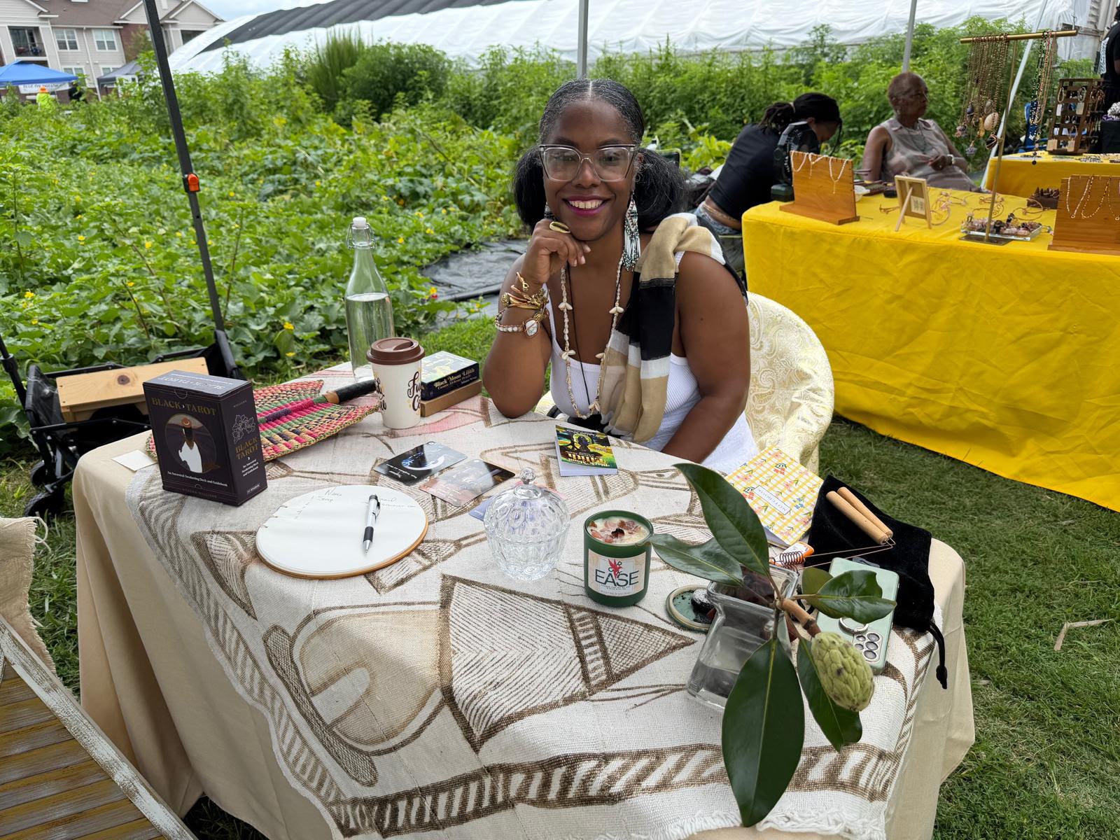 Smiling woman sitting at a table outdoors with various items, greenery in background, and a yellow tablecloth nearby.