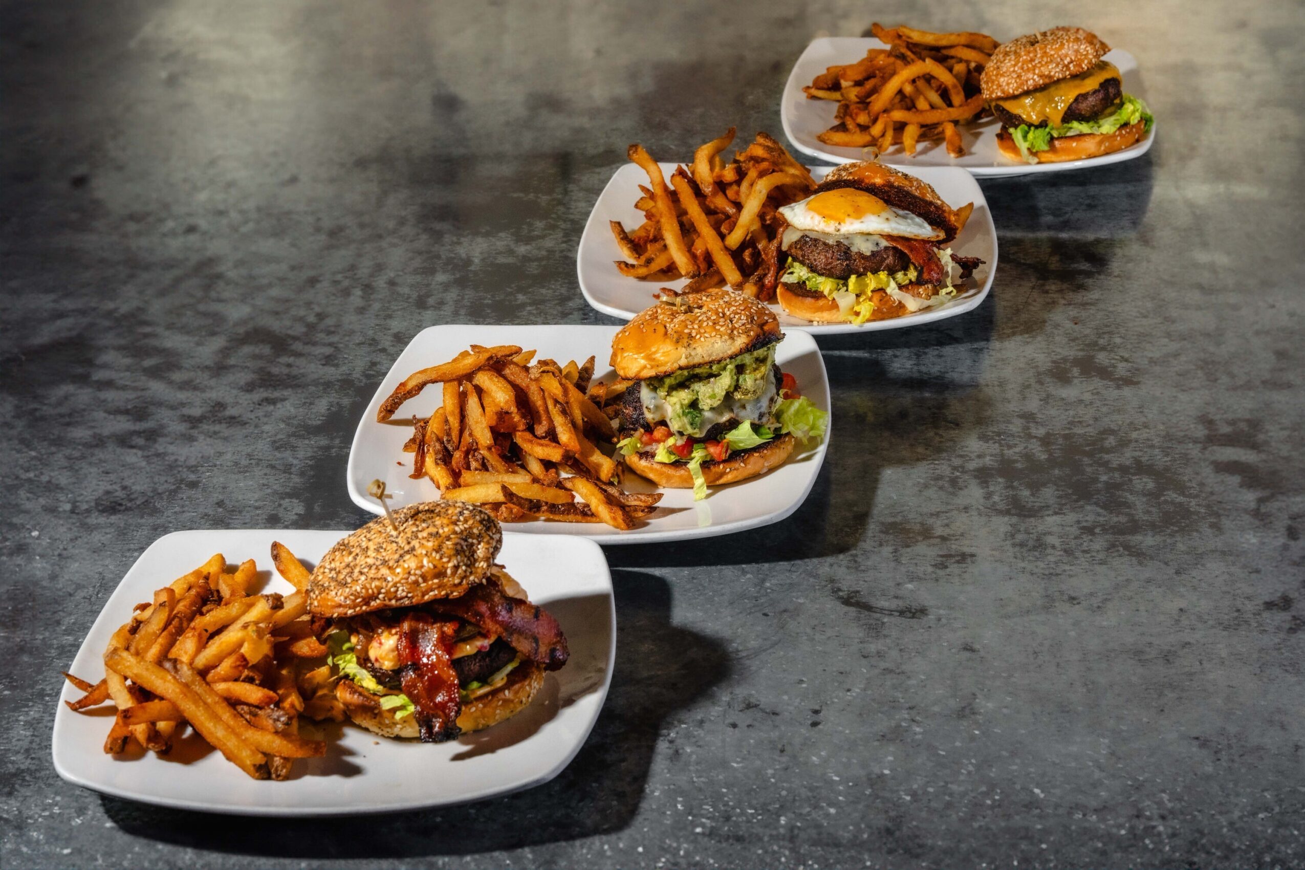 Four plates of burgers and fries arranged diagonally on a dark tabletop.