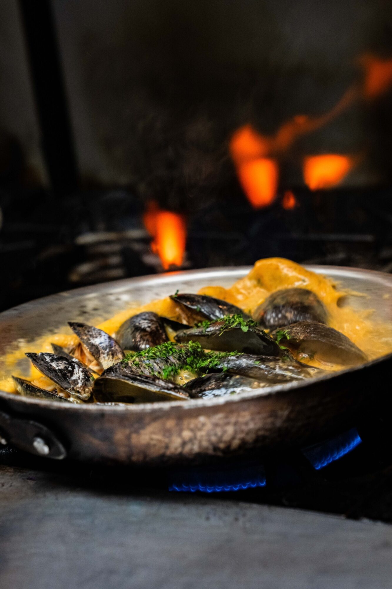A pan of mussels in sauce sits over a blue gas flame, with flames visible in the background.