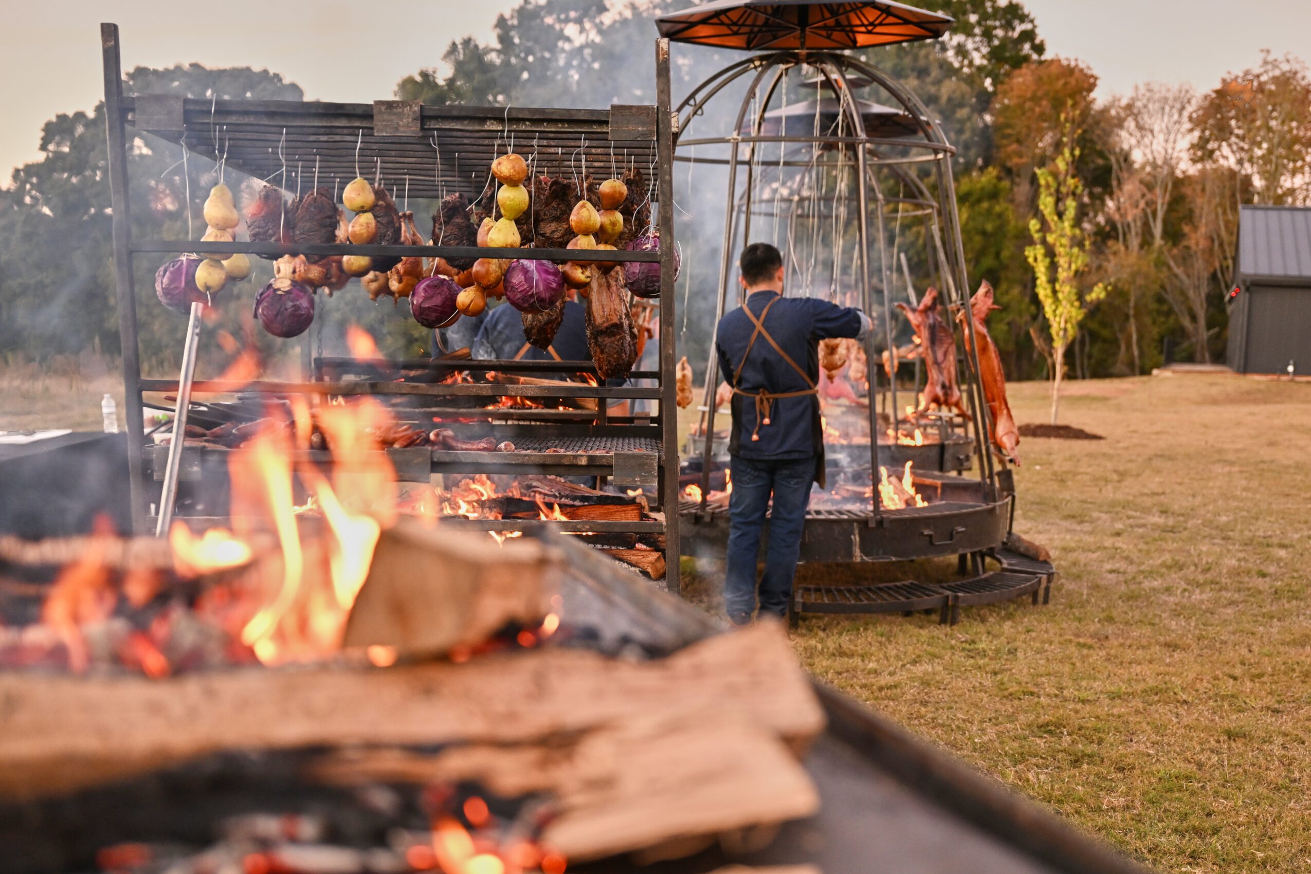 Person tending a large outdoor grill with hanging vegetables and open flames in a grassy area.