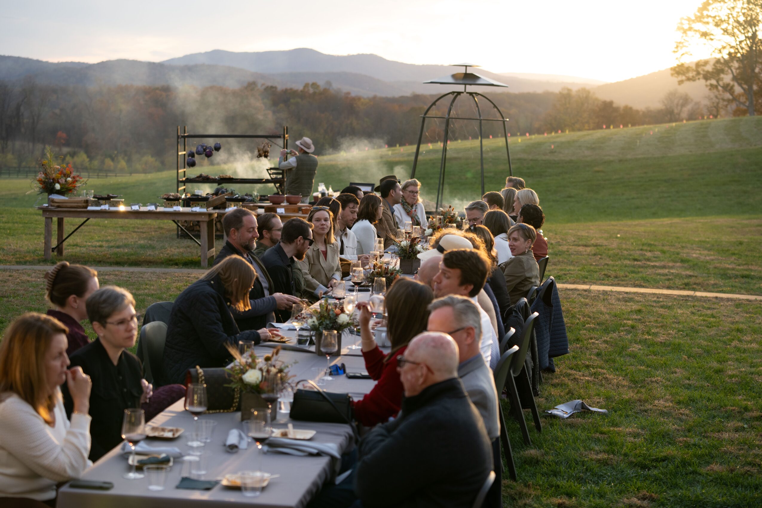 People seated at a long outdoor dinner table in a grassy field, with hills and trees in the background.
