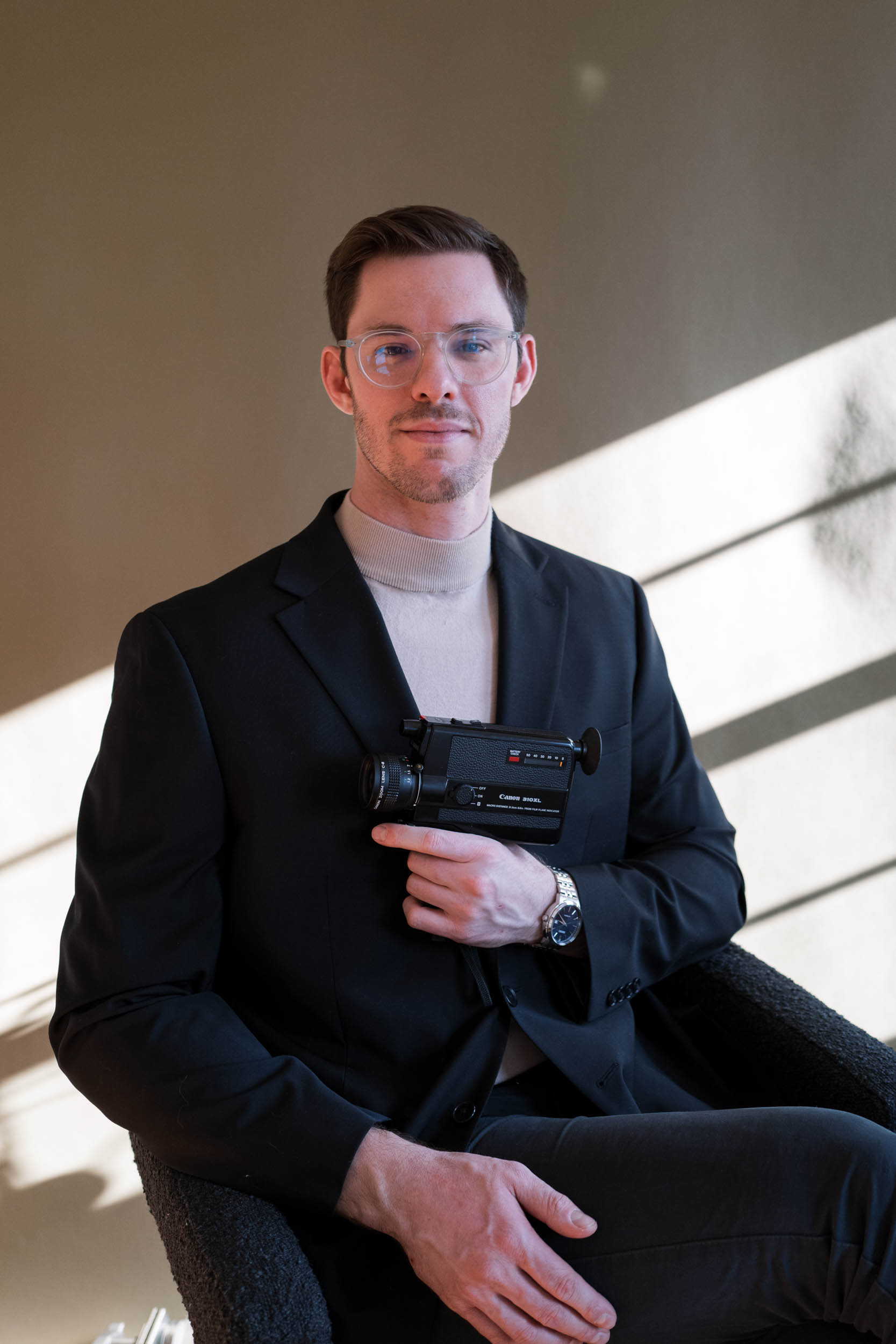 Man in black suit holding a video camera, sitting indoors with sunlight and shadows, neutral expression.