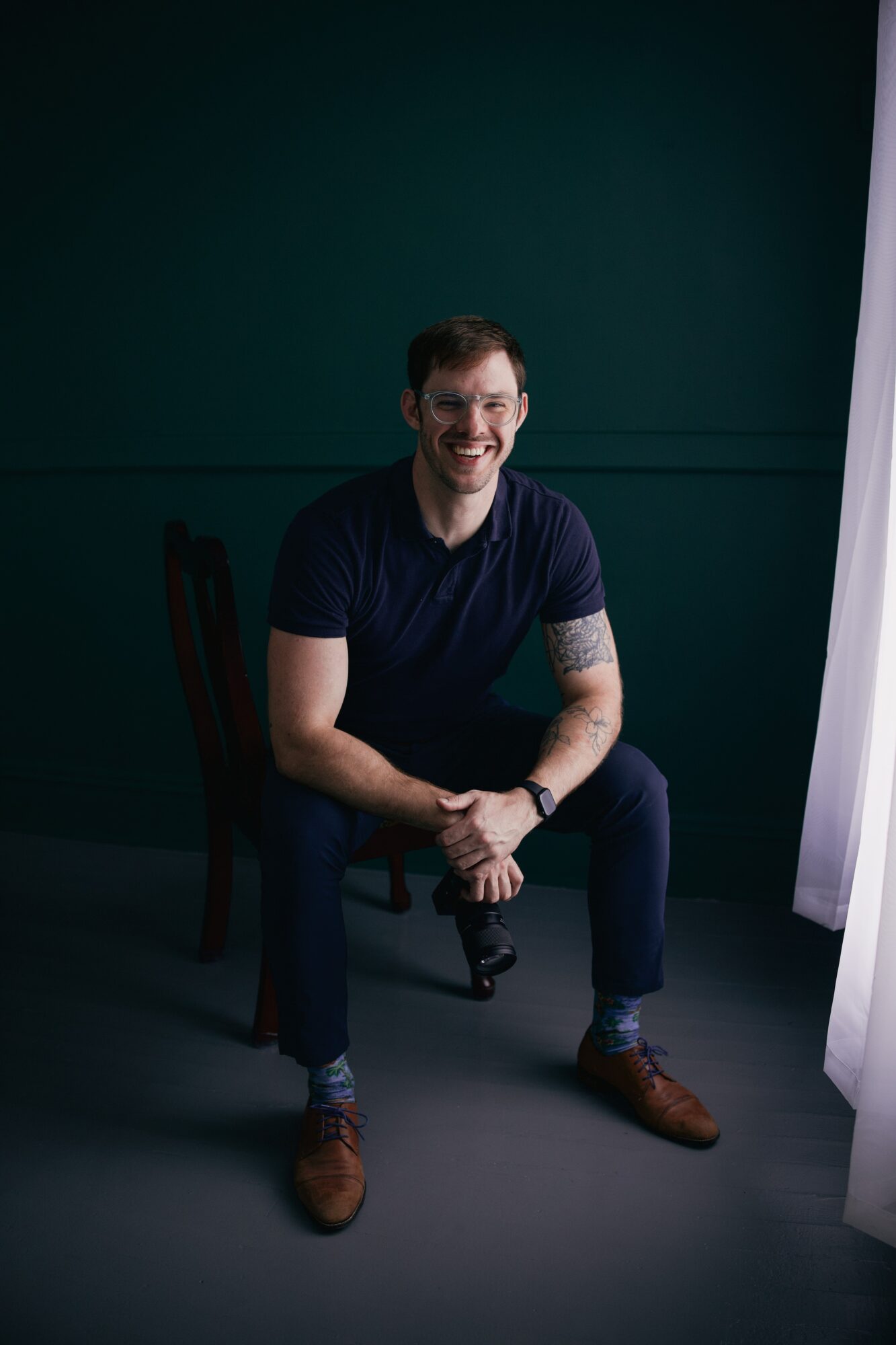 Young man with short hair and beard, sitting on a chair, smiling, wearing a dark t-shirt and jeans, in a studio setting.