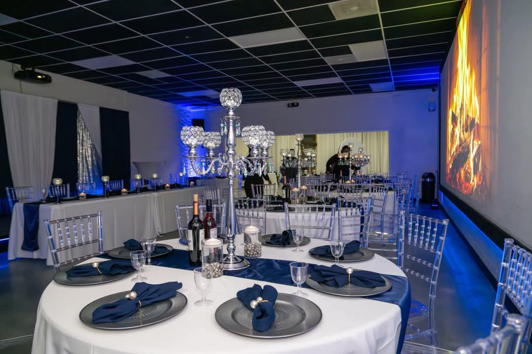 Elegant event space with round tables, blue napkins, a chandelier, and a large screen displaying a fiery image.