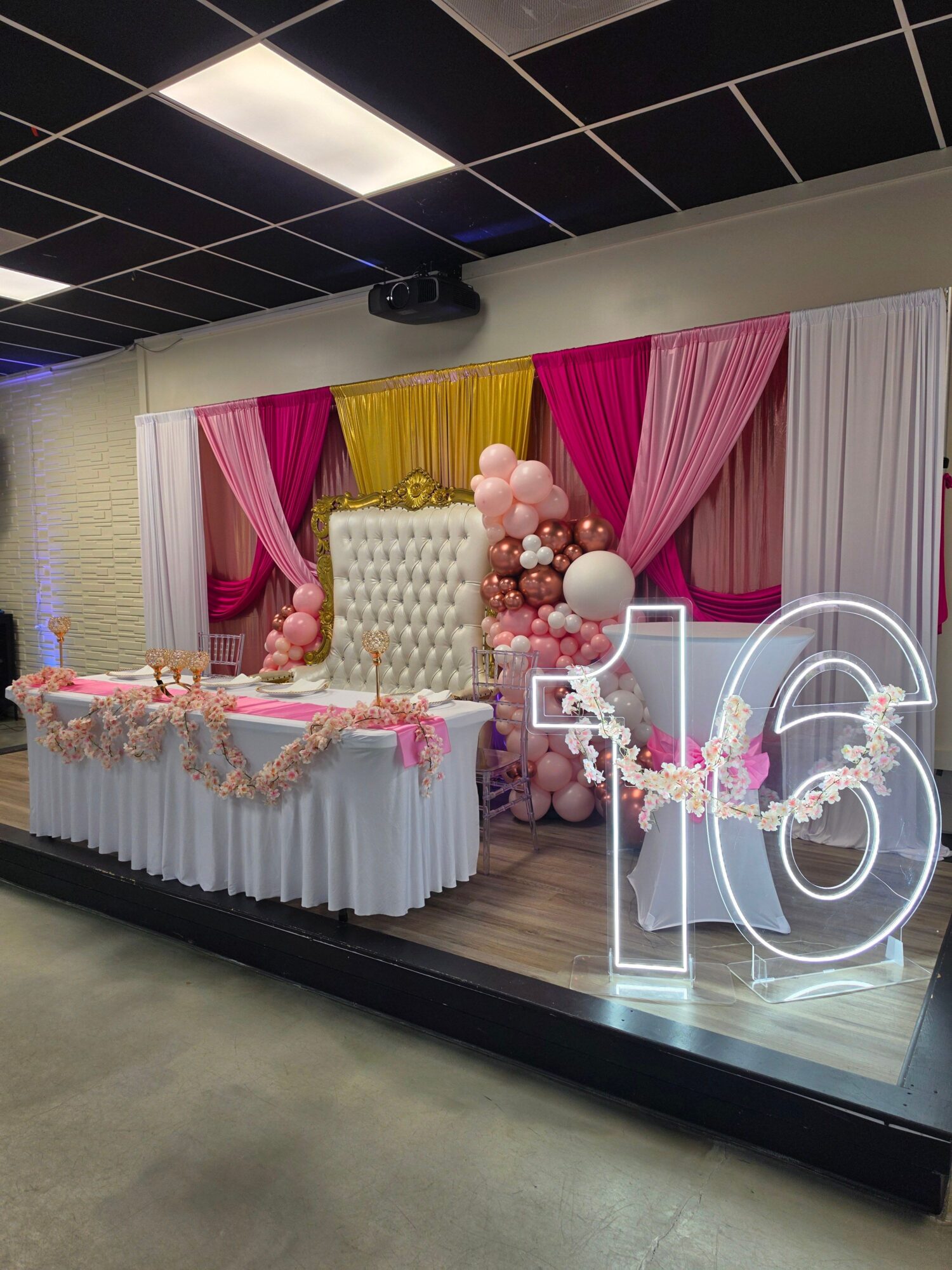 Decorative table with pink, yellow, and white drapes, a large illuminated number 2, and floral arrangements for celebration.