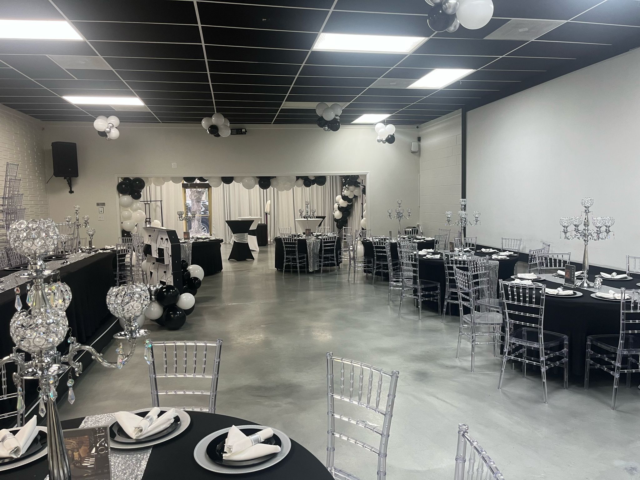 Event hall with decorated tables, chairs, and chandeliers, prepared for a celebration or gathering, with a white wall and black ceiling.