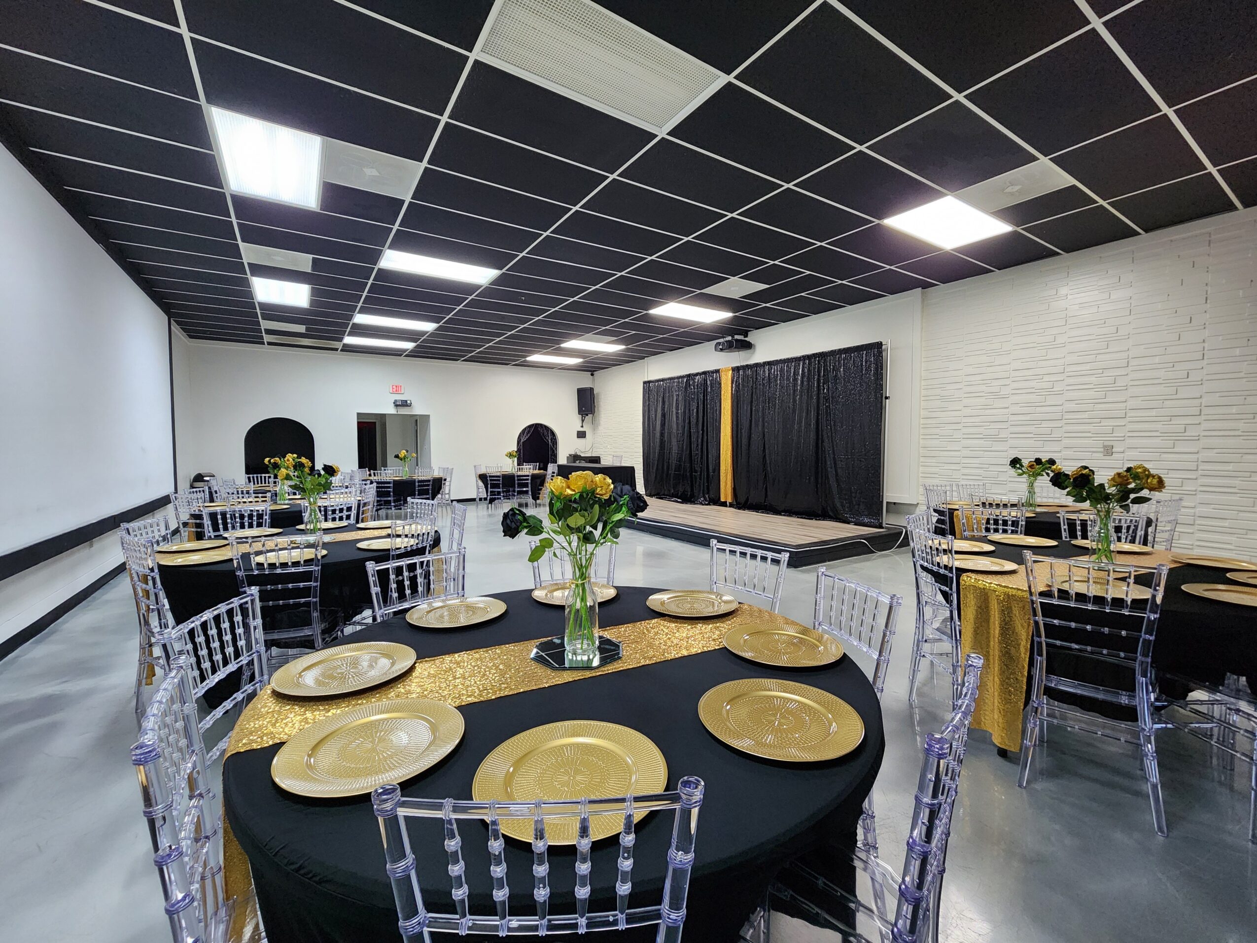 Event space with round tables, black tablecloths, clear chairs, and a stage with black curtains, decorated with flowers.
