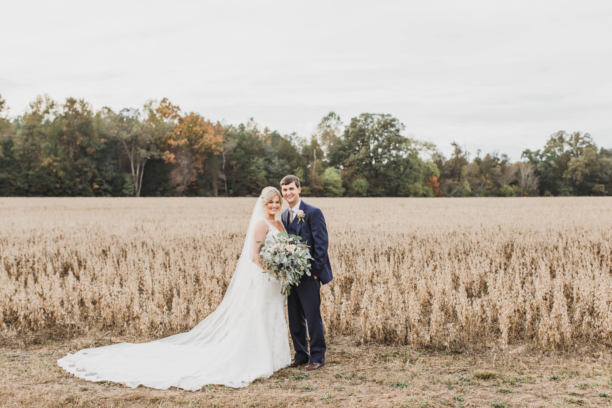 A bride and groom stand together in a field, smiling, with trees in the background, bride holding a large bouquet, wedding attire.