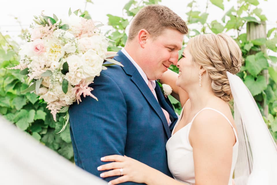 A bride and groom face each other closely, smiling, with greenery and flowers in the background.