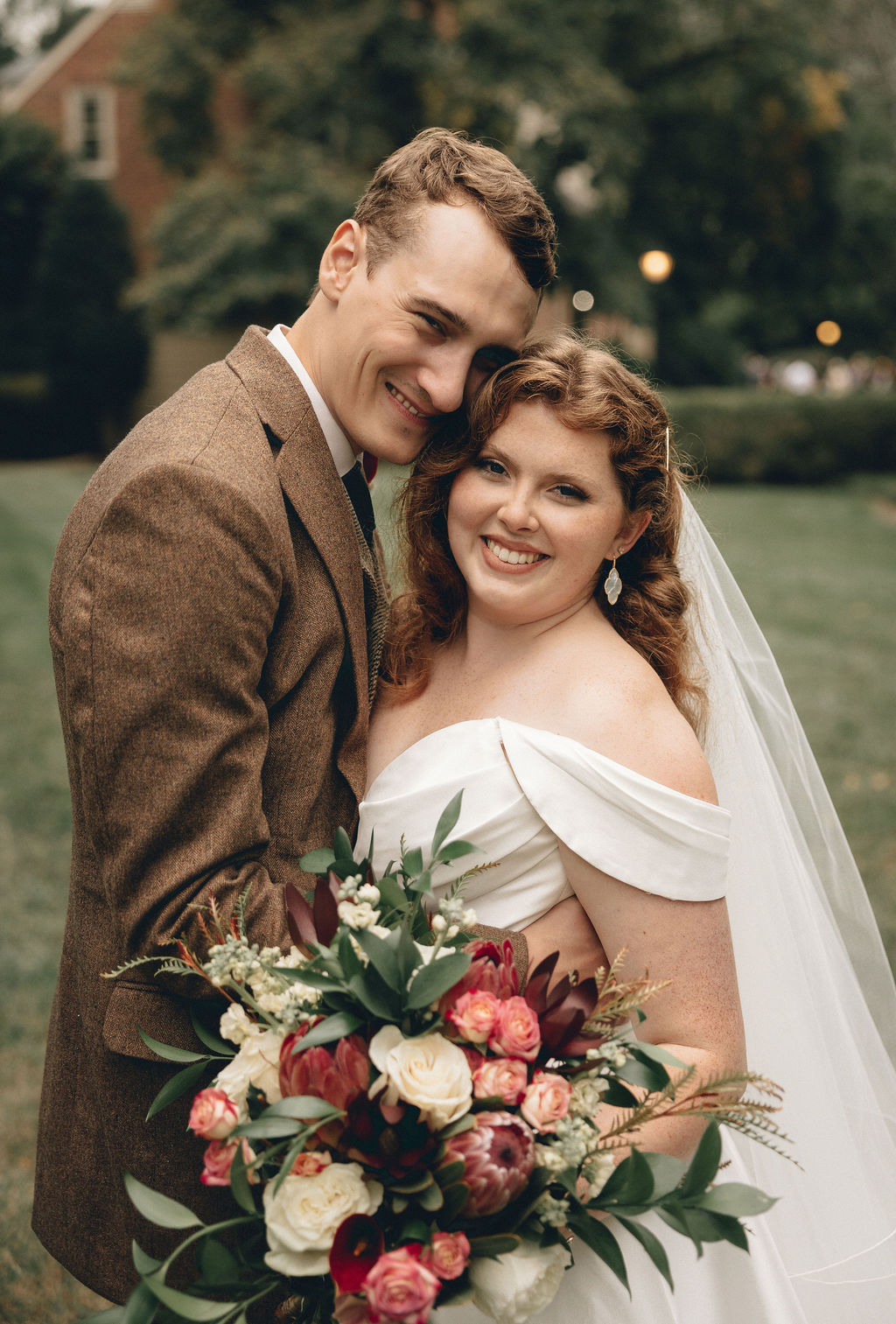 A smiling bride and groom embrace outdoors, holding a bouquet of flowers, with trees and buildings in the background.
