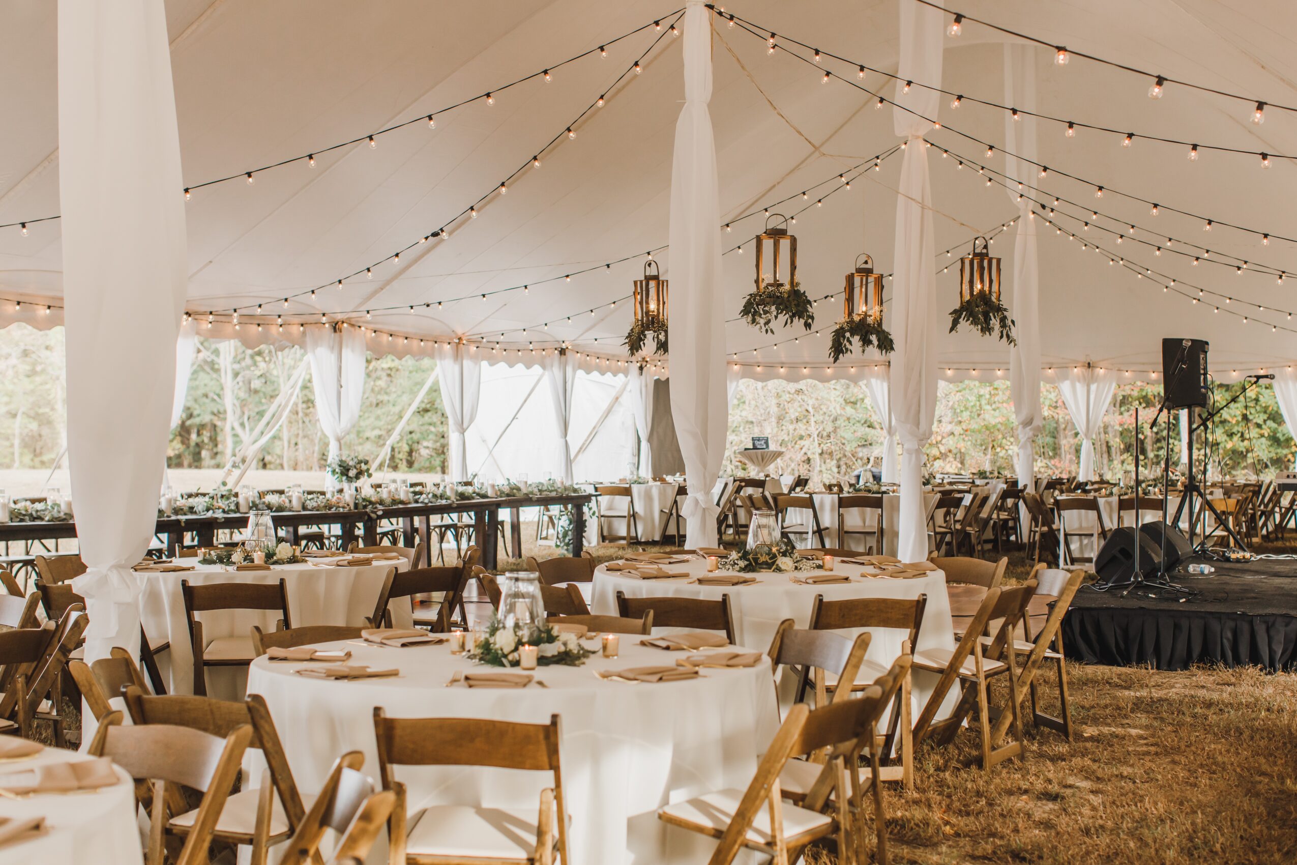 Event tent with round tables, chairs, string lights, and decorative lanterns, set up for a celebration or gathering.