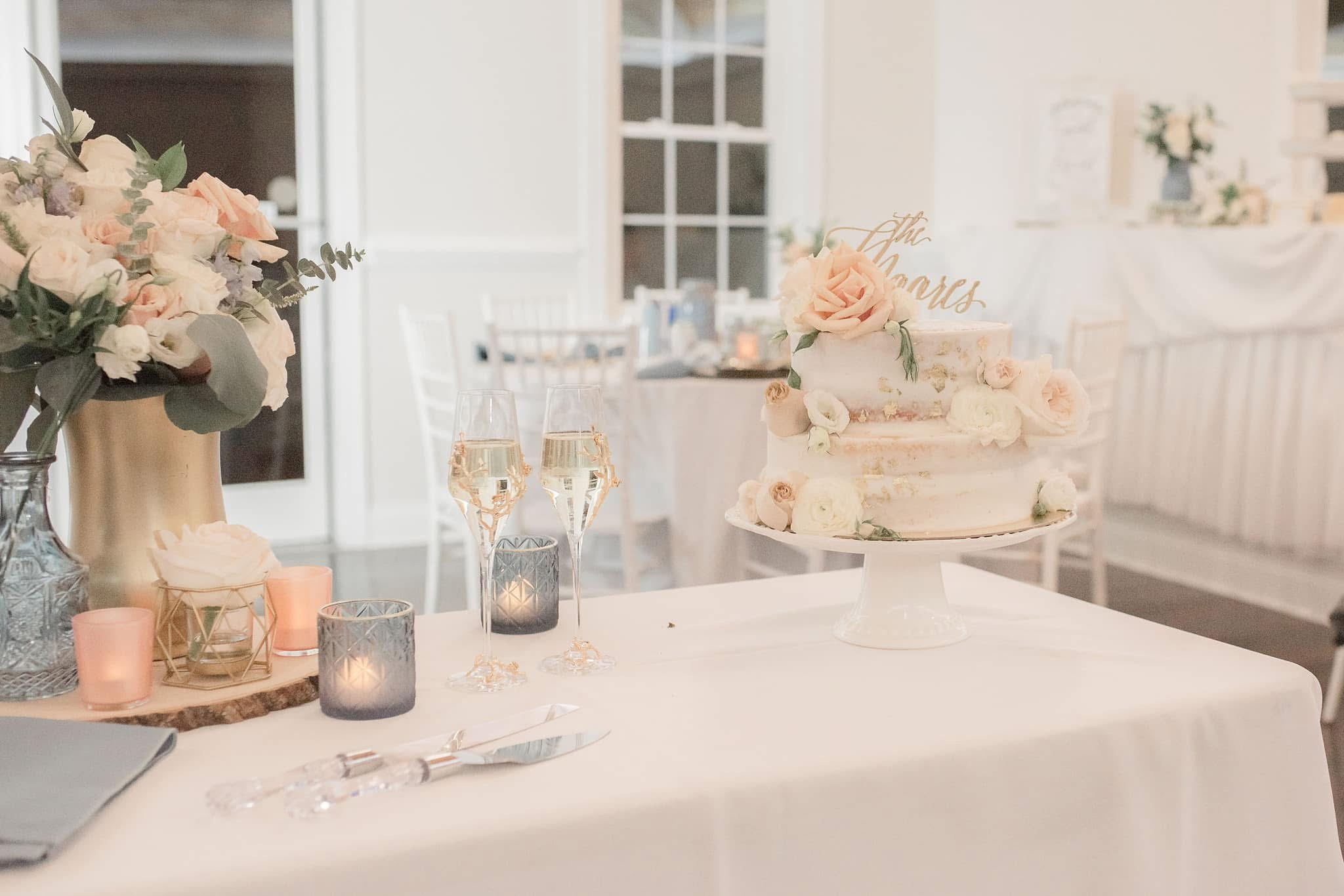 Wedding reception table with floral centerpiece, cake, candles, and glasses, in a bright, elegant room.