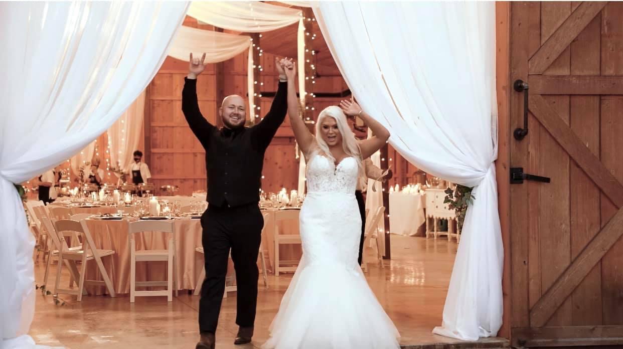 A bride and groom celebrate at their wedding reception, holding hands and raising arms, with decorated venue in background.
