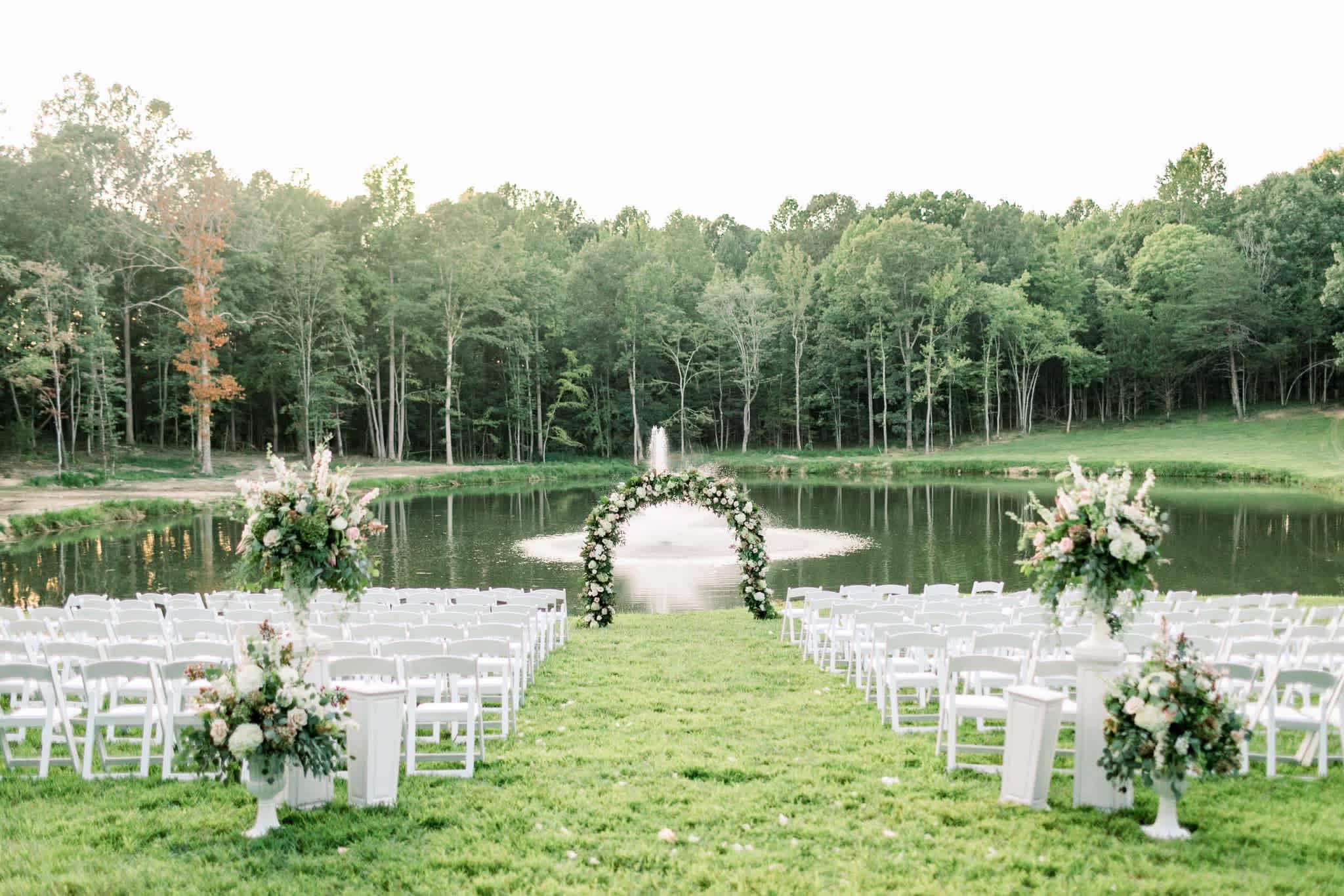 Outdoor wedding setup with chairs facing a floral arch by a lake, surrounded by trees, under a cloudy sky.