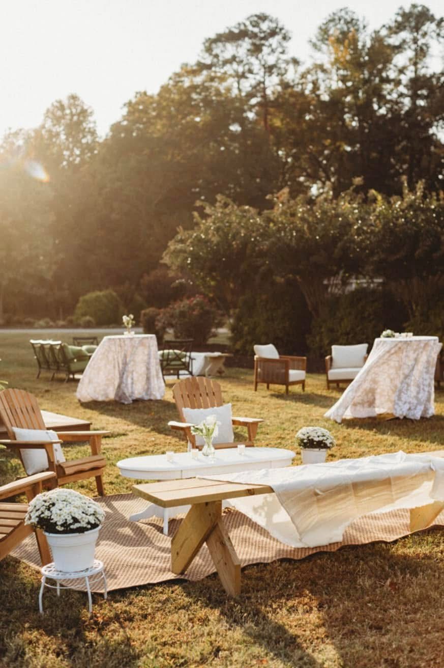 Outdoor setting with tables and chairs on grass, surrounded by trees, sunlight, and floral decorations.