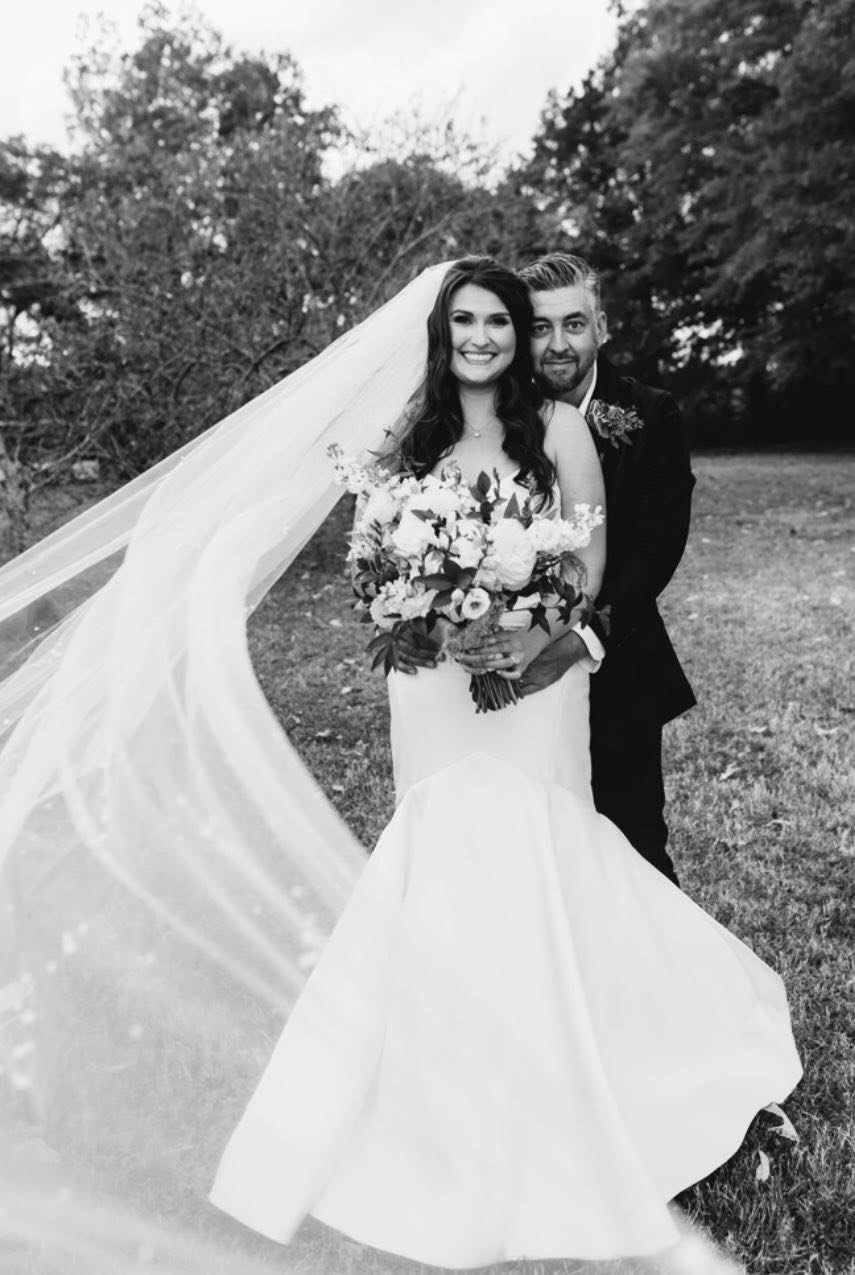 A bride and groom outdoors, smiling, with trees in the background. The bride wears a wedding dress with a long veil, holding a bouquet. The groom is in a suit.