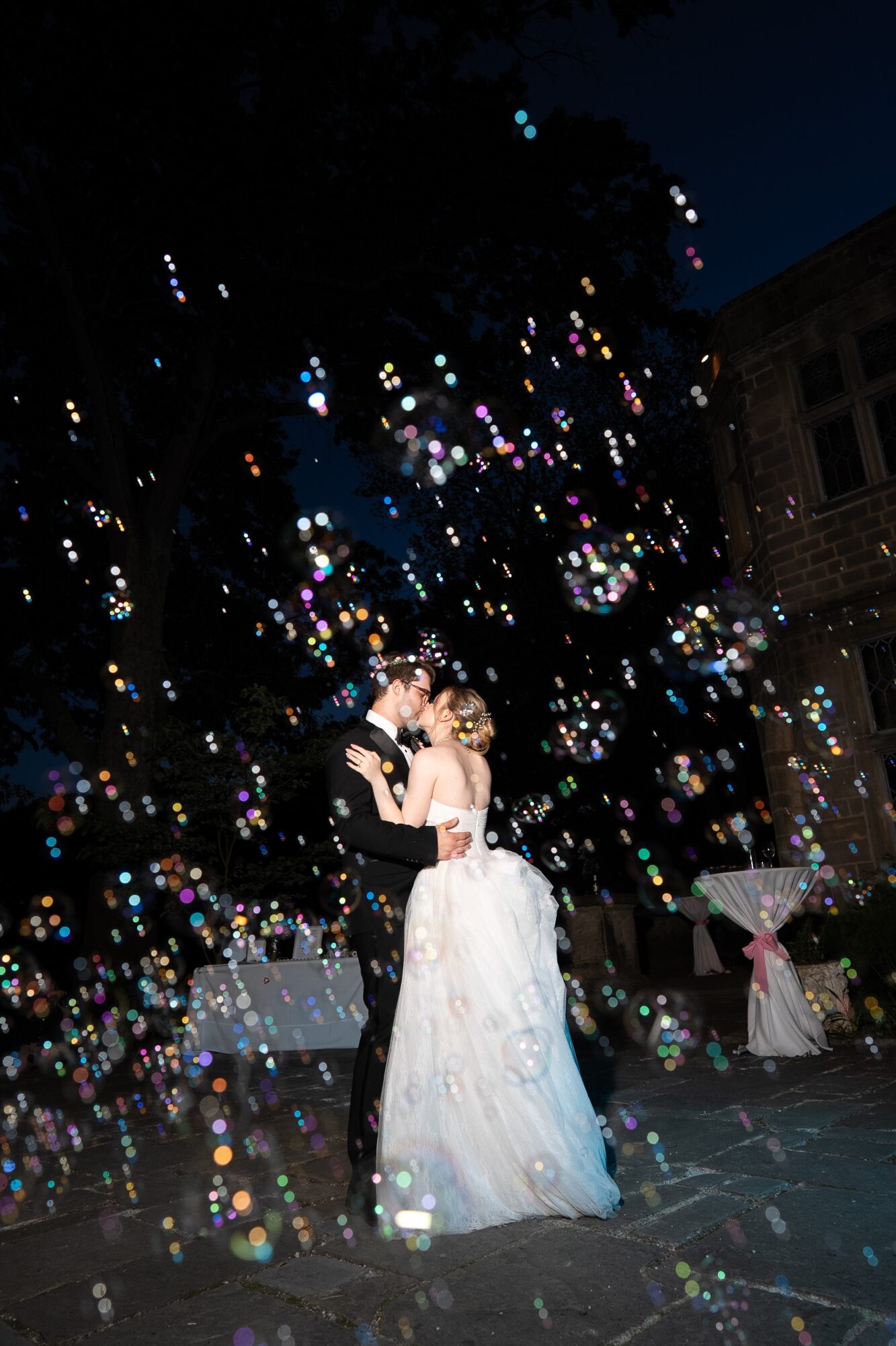 A couple in wedding attire dancing outdoors at night surrounded by colorful bubbles and illuminated by lights.