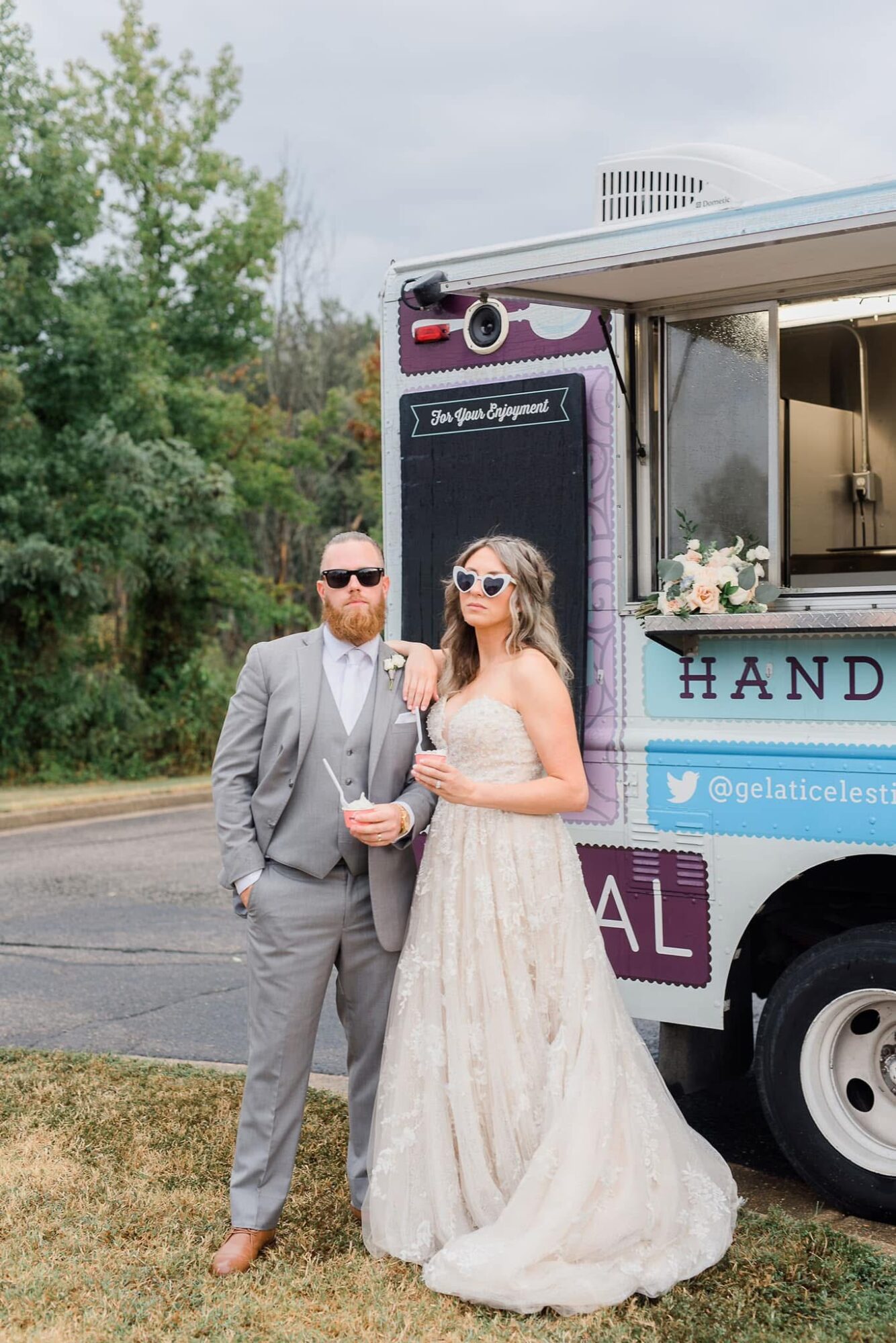 A couple in formal attire standing next to a food truck outdoors, with trees and cloudy sky in background.