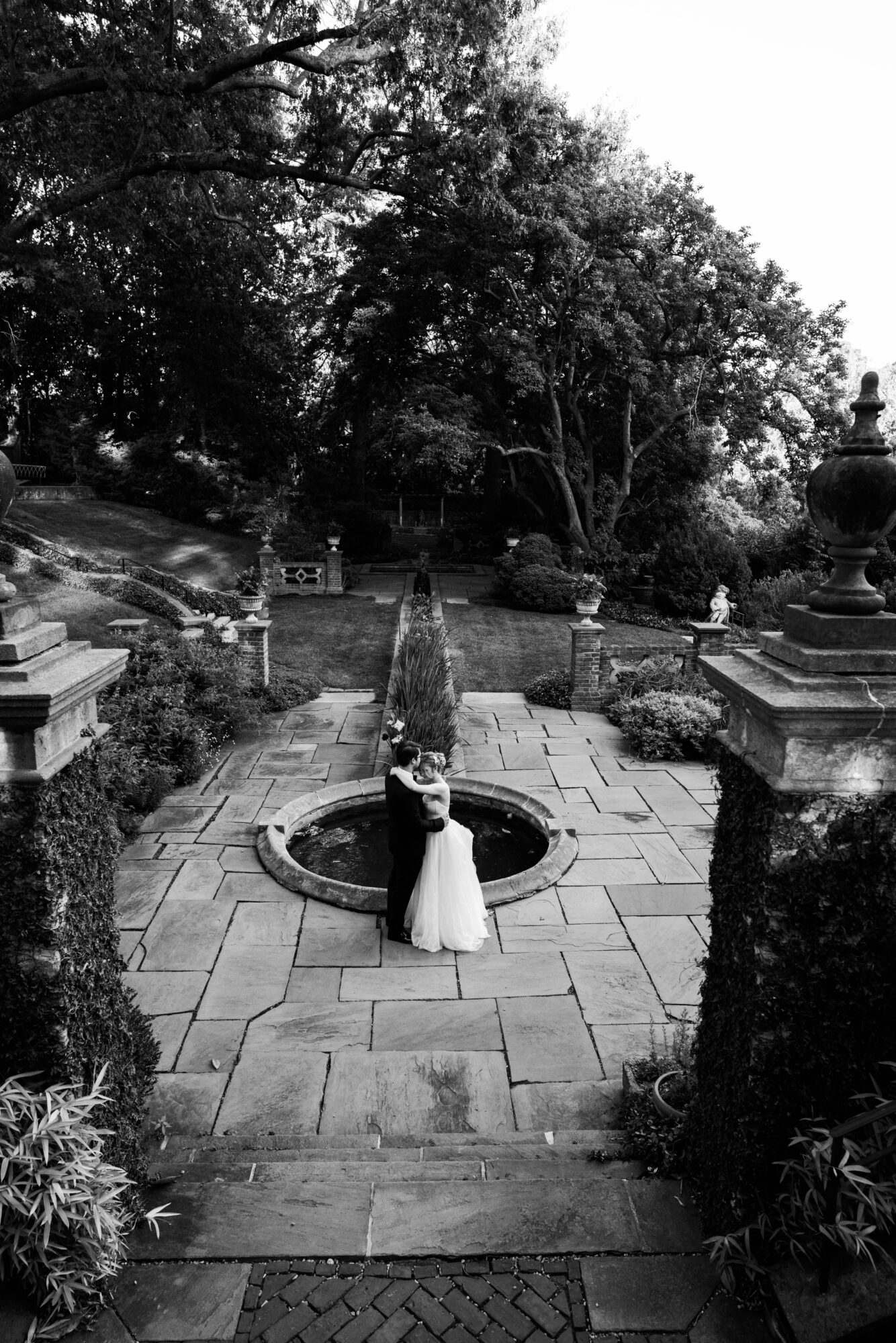 A bride and groom stand together in a garden with trees and stone structures, holding hands near a circular fountain.
