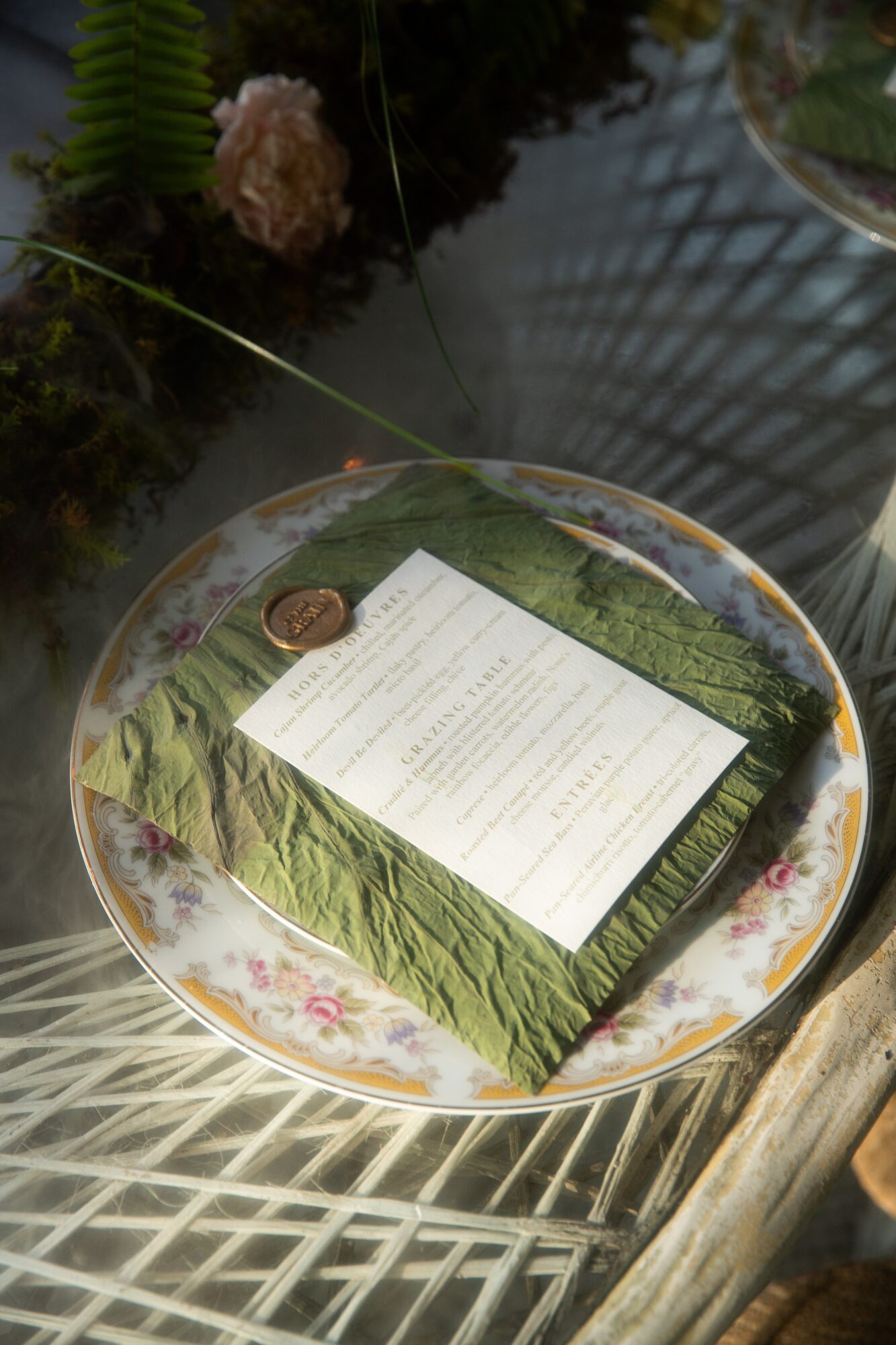 Book with a green cover and a sheet of paper on top, placed on a floral-patterned plate, with plants and a glass surface nearby.