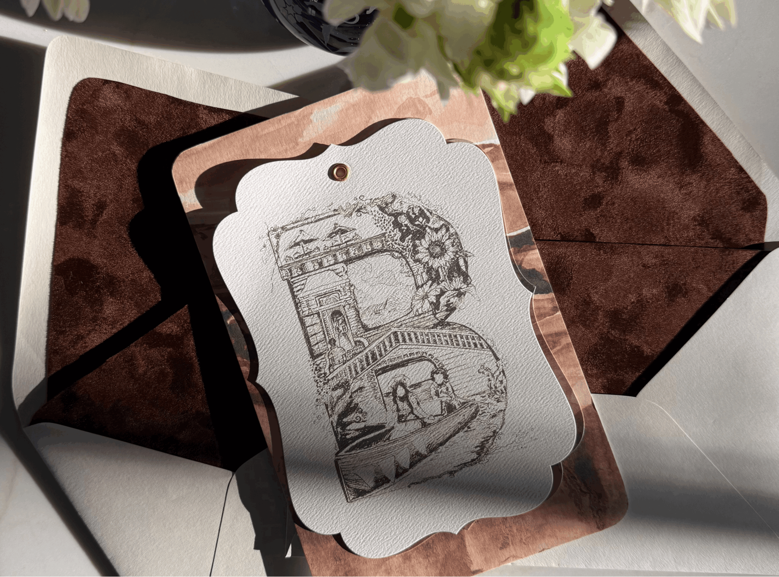 Decorative card with sketches of a building and a bridge, placed on a brown surface with flowers above.
