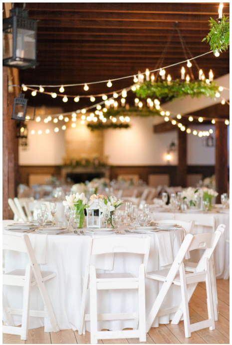 Event space with round tables, white chairs, string lights, and greenery, decorated for a celebration or wedding reception.
