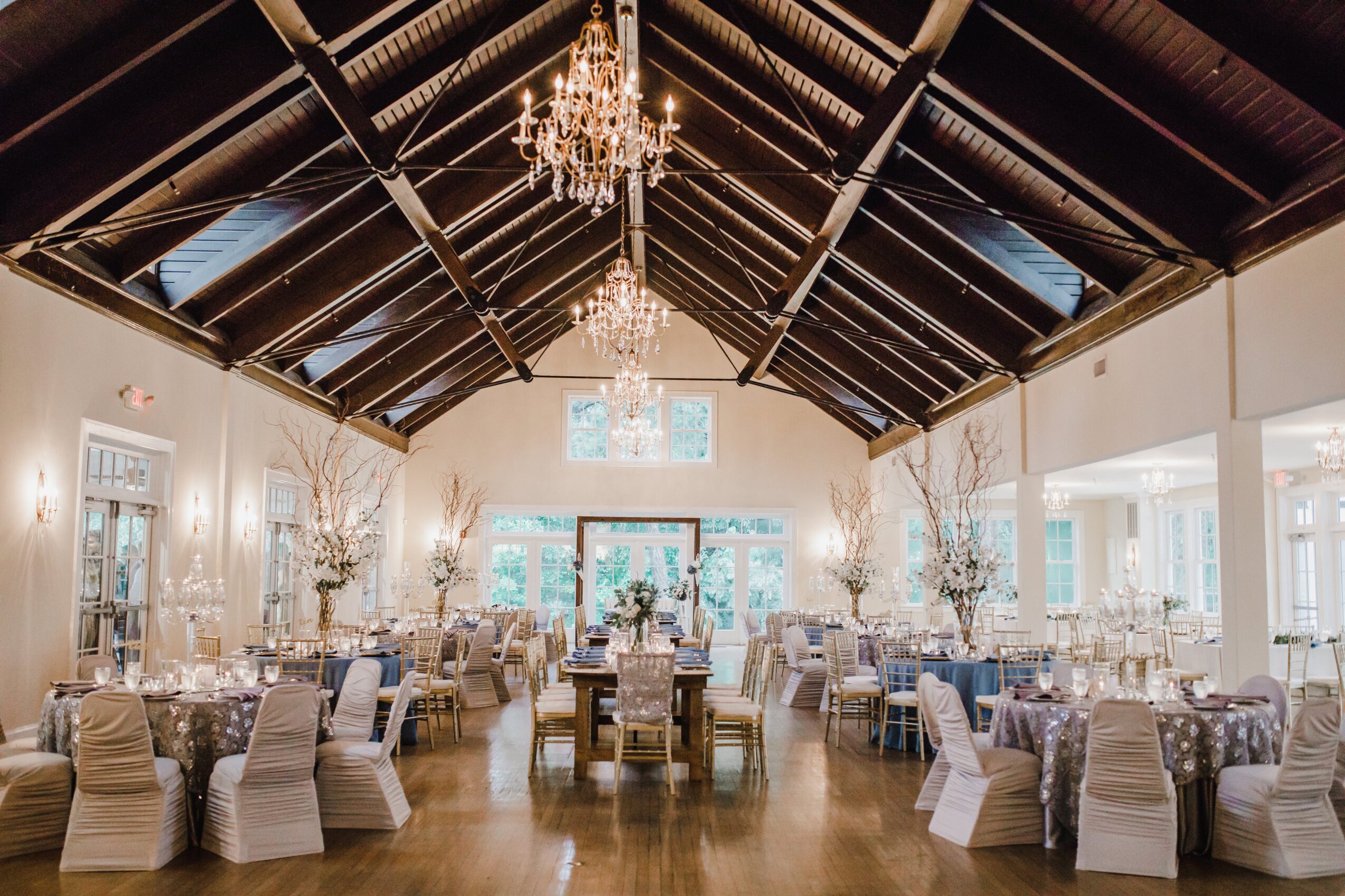 Elegant banquet hall with chandeliers, decorated tables, and large windows, featuring a high vaulted ceiling with wooden beams.