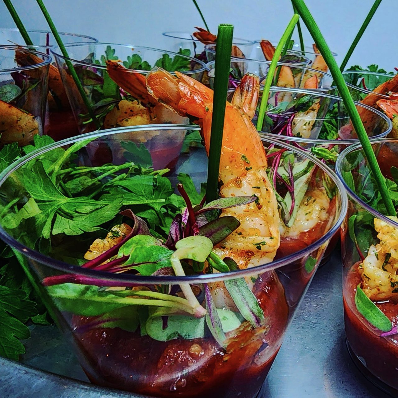 Close-up of shrimp and mixed greens in a glass cup with a skewer, fresh herbs, and vegetables.