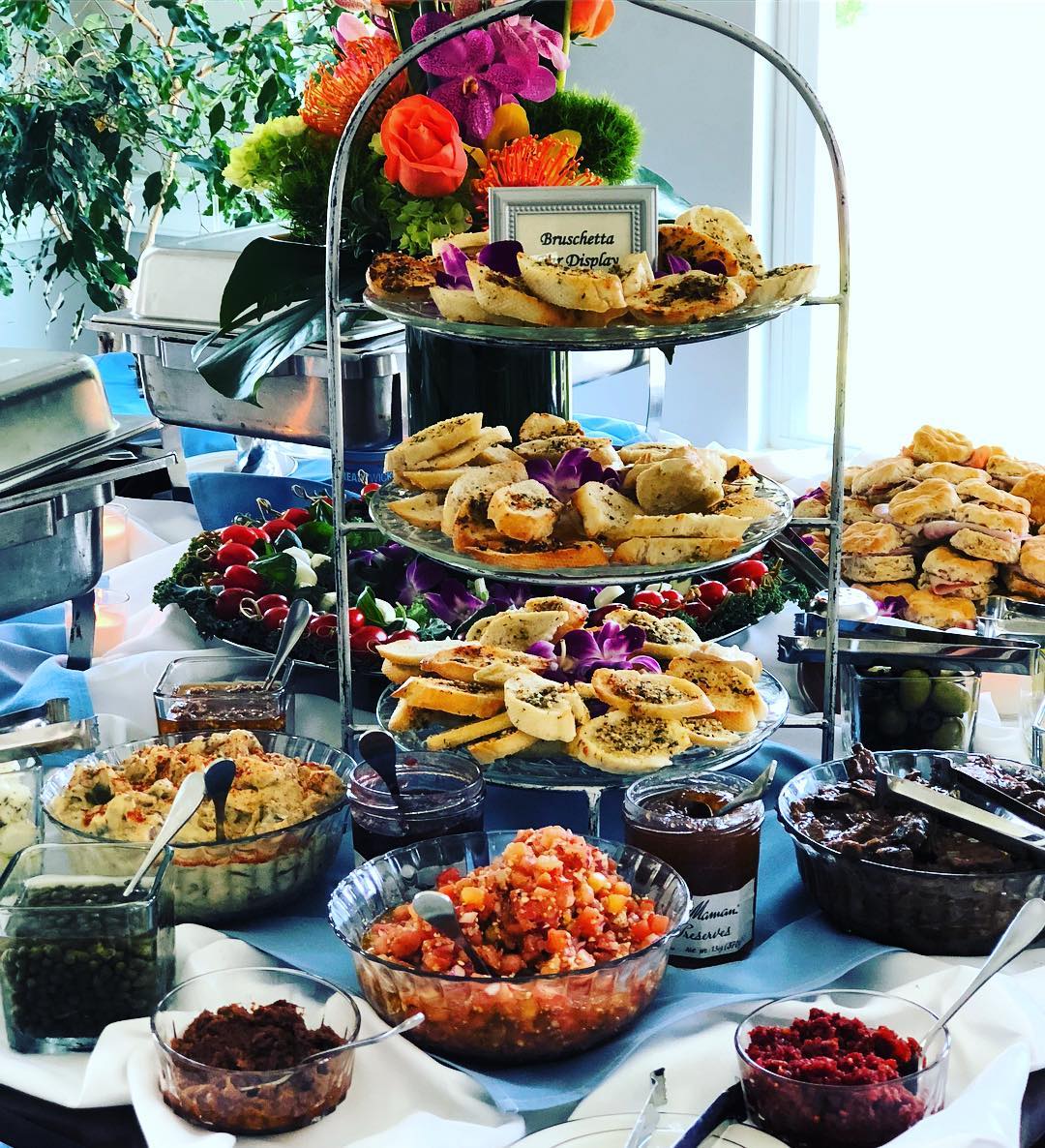 Three-tiered tray with assorted cookies, pastries, and a small sign, surrounded by bowls of fruit and jams, with flowers in background.
