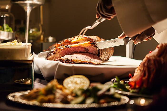 Person carving a large roasted meat on a white platter, with food and drinks on a table, in a dimly lit setting.