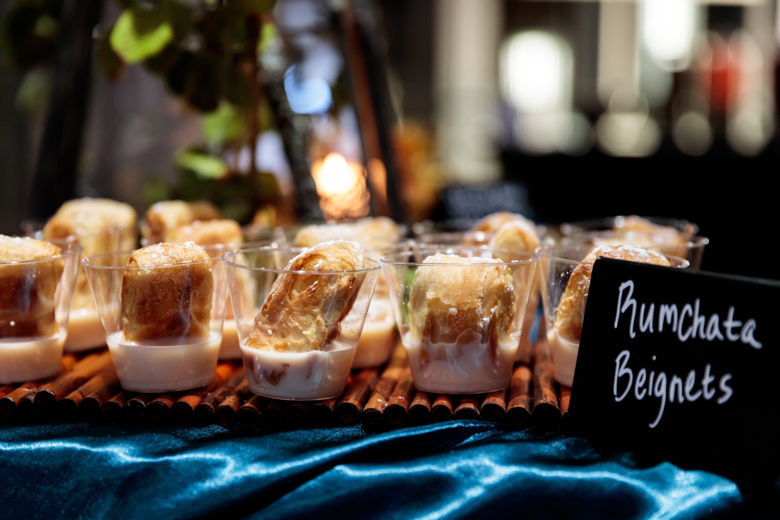 Small glasses with Rumchata and beignets on a table, with a sign reading 'Rumchata Beignets'.