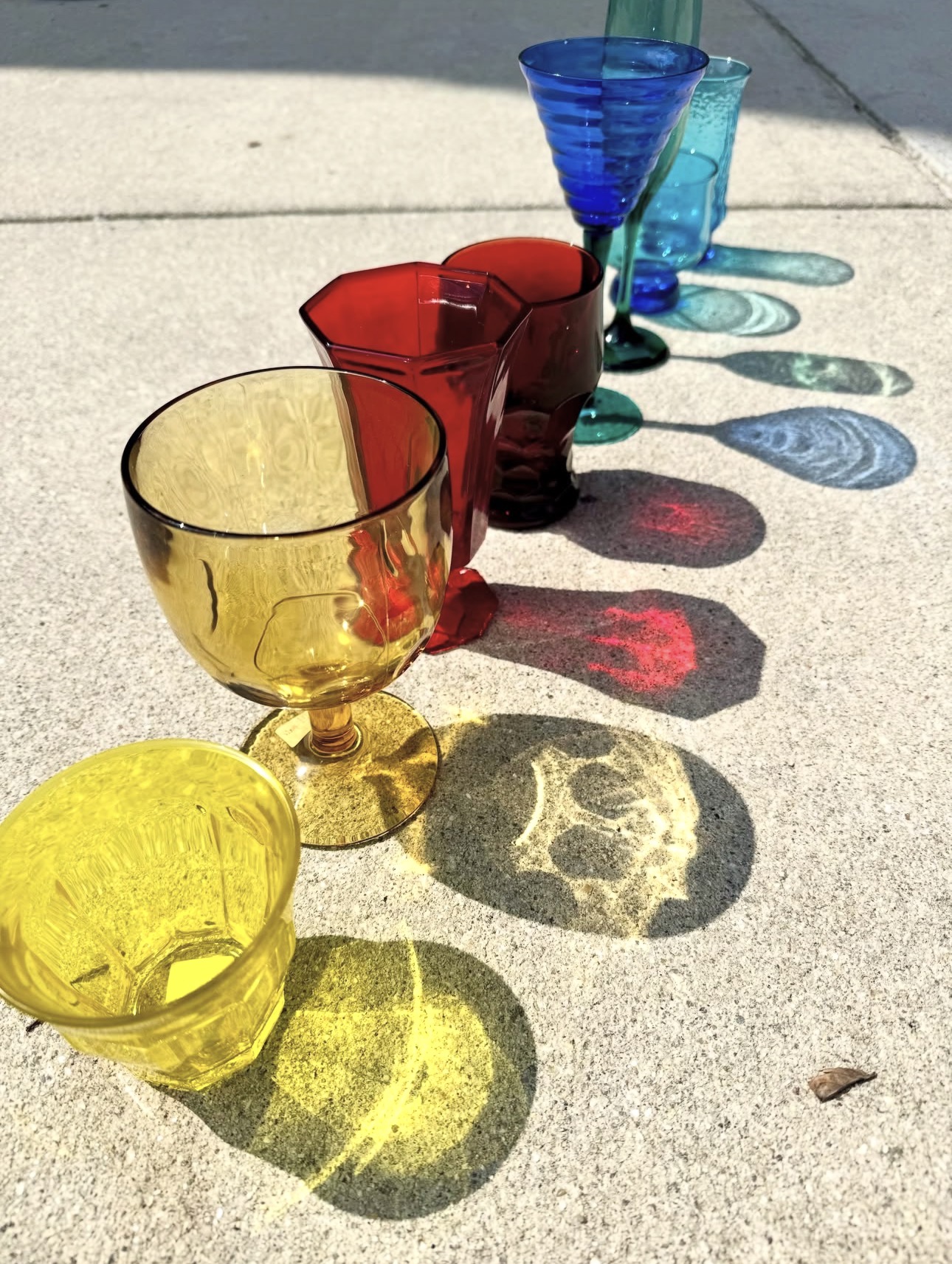 Colorful glass cups arranged in a line on a concrete surface, casting shadows in sunlight.
