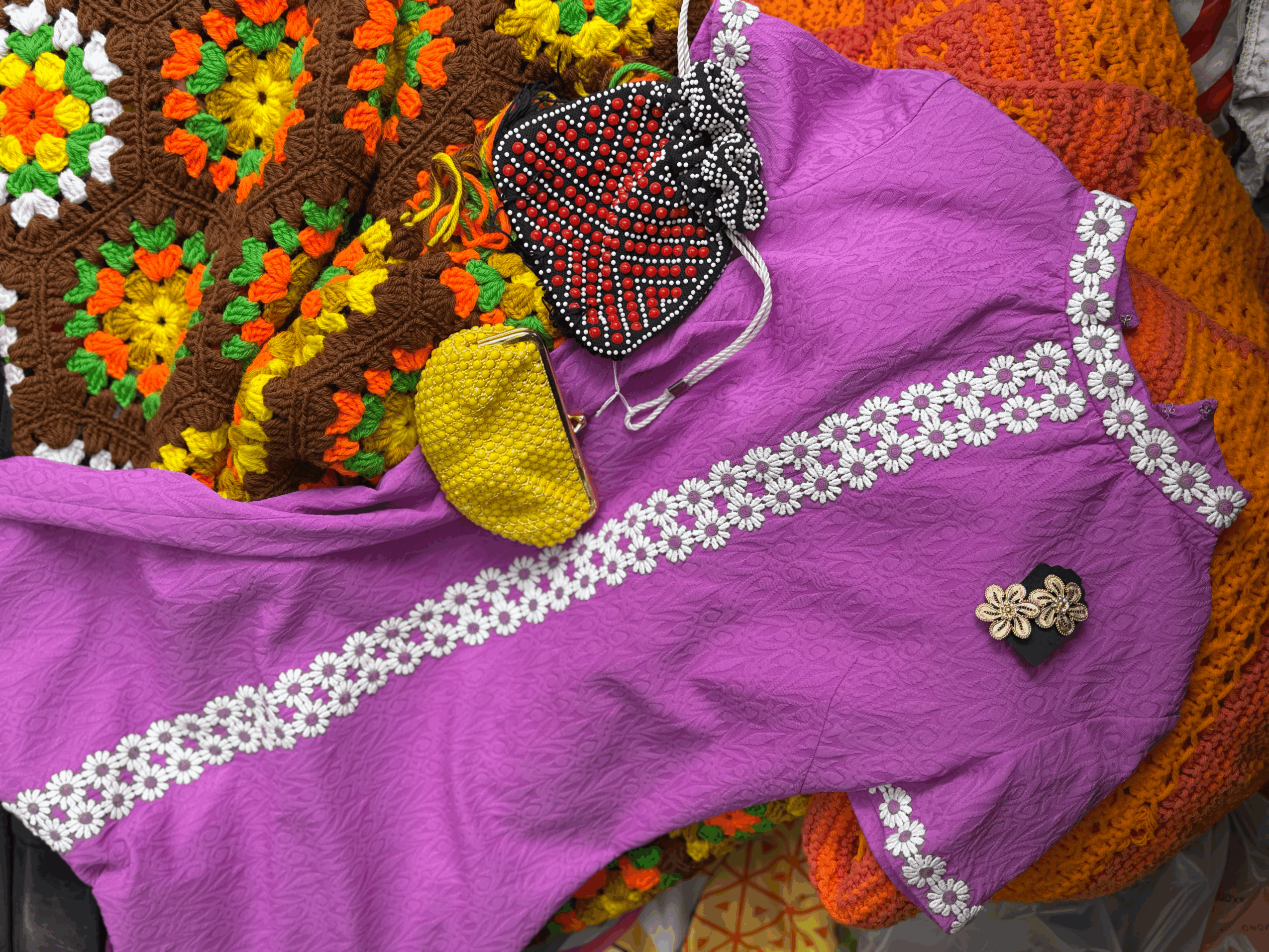 Colorful handmade crochet items and a purple fabric with white floral trim, laid on a multicolored textured background.