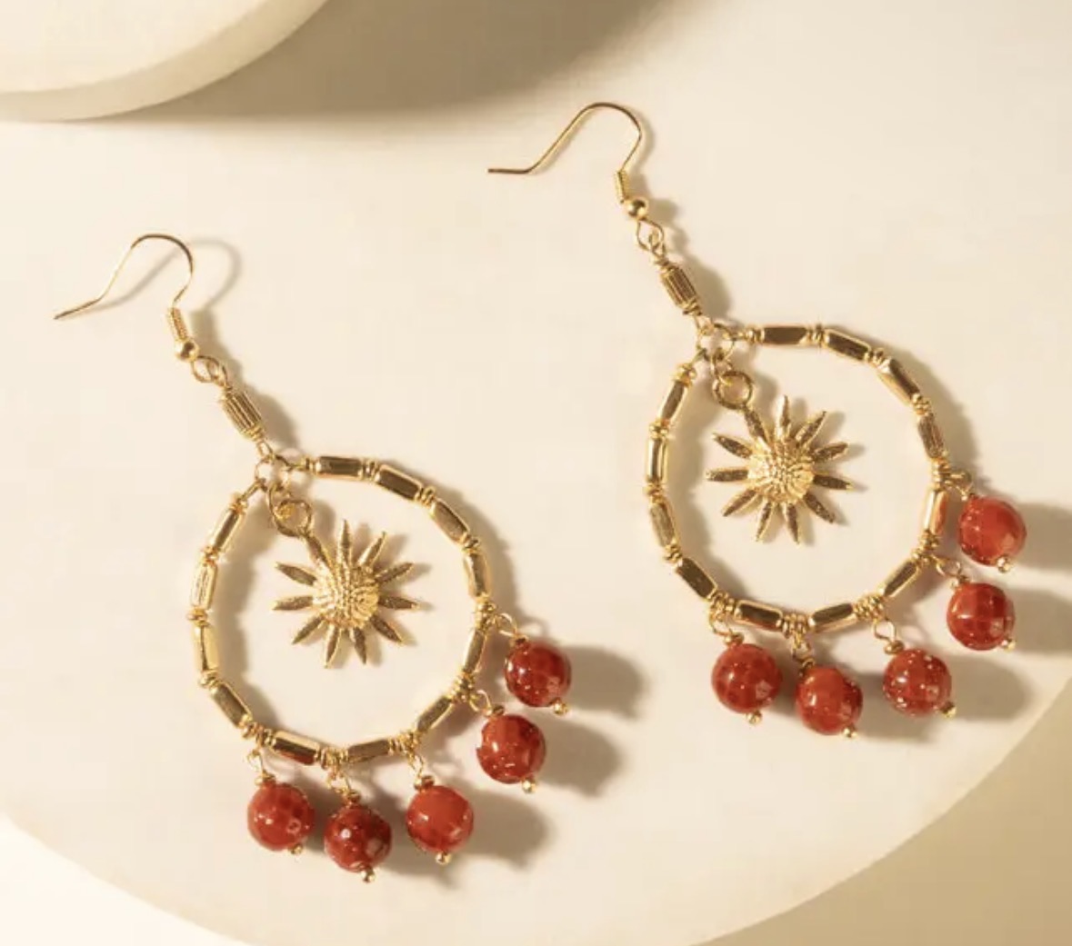 Pair of gold hoop earrings with sunburst charms and red bead dangles on a cream surface.