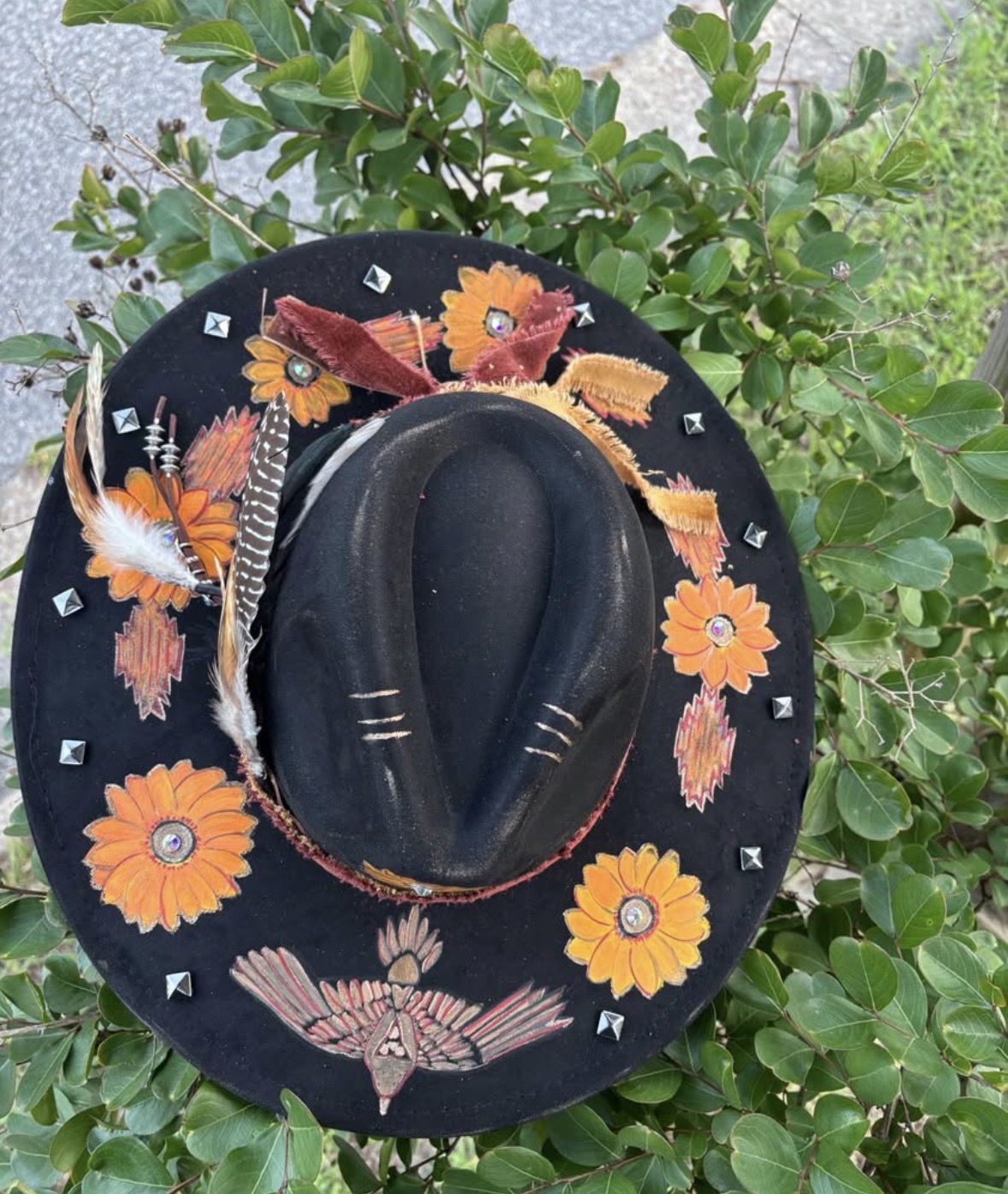 Black wide-brimmed hat decorated with orange flowers and feathers rests among green leaves.