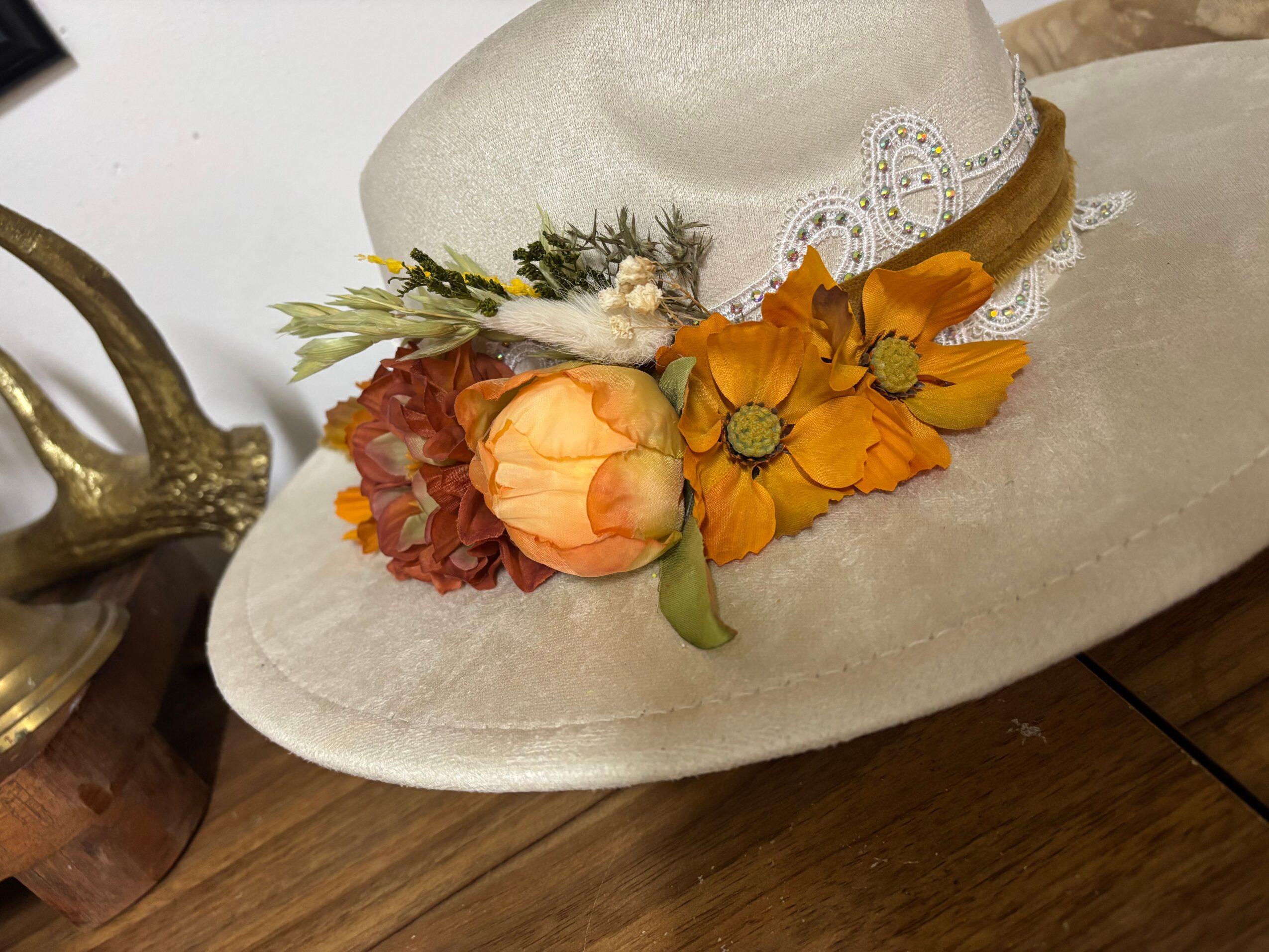 Cream-colored hat decorated with orange flowers, leaves, and a jeweled band rests on a wooden floor.