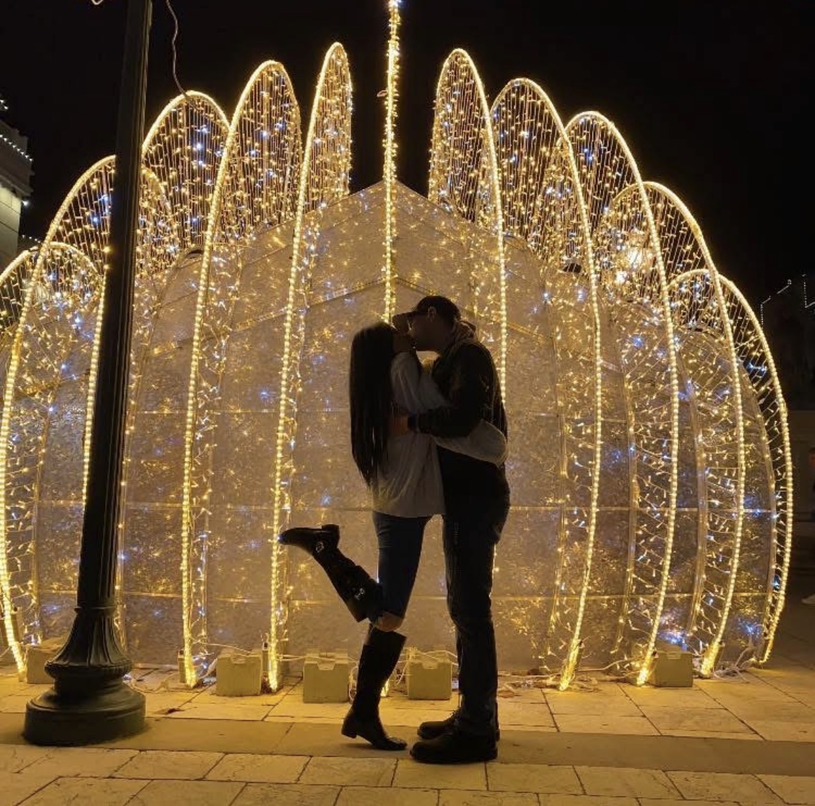 Two people kiss beneath a large illuminated arch decorated with golden lights at night.