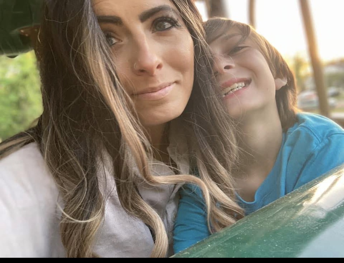 Two people pose closely for a selfie outdoors, with greenery and a bright window behind them.