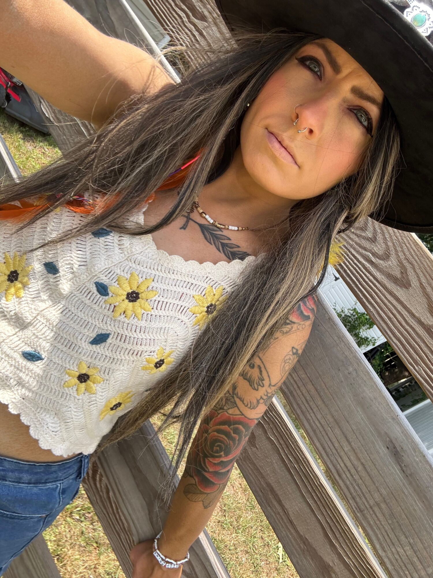 A person with long dark hair sits on a wooden bench, wearing a white floral crochet top and displaying arm tattoos.