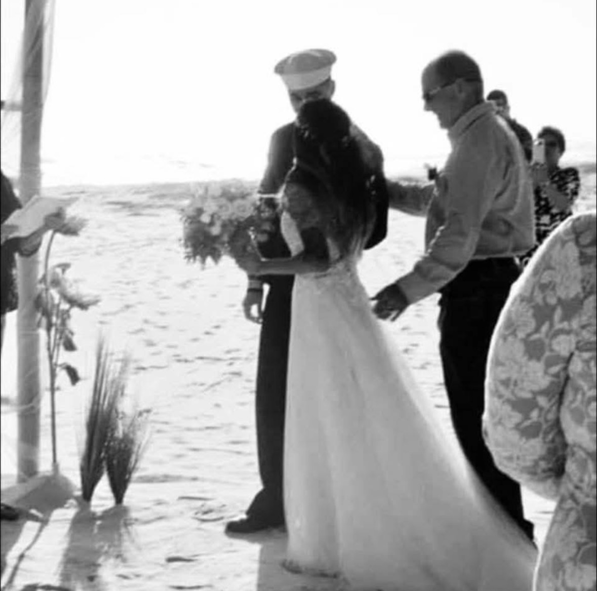 A person in a long white dress holds a bouquet beside another person on a beach, with guests nearby.