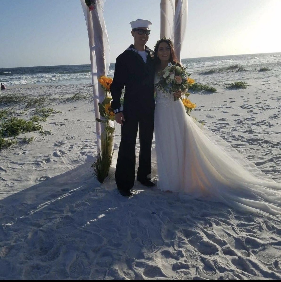 A couple stands beneath a decorated beach arch, with ocean waves and sand behind them.