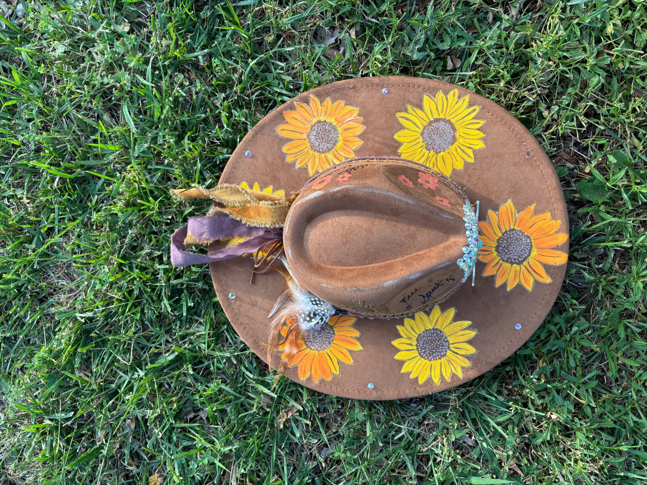 A brown cowboy hat decorated with painted sunflowers rests on green grass.