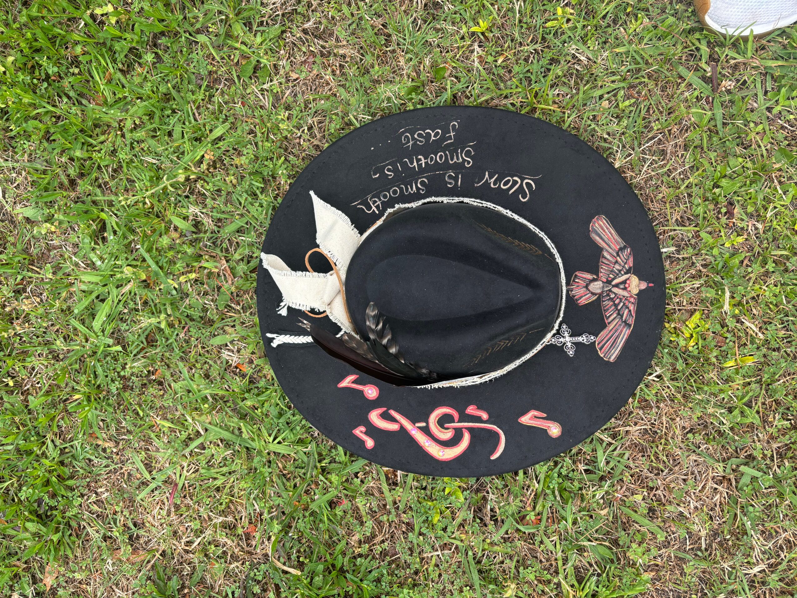 Black decorated hat with a central oval opening lies on grass beside a small white object.