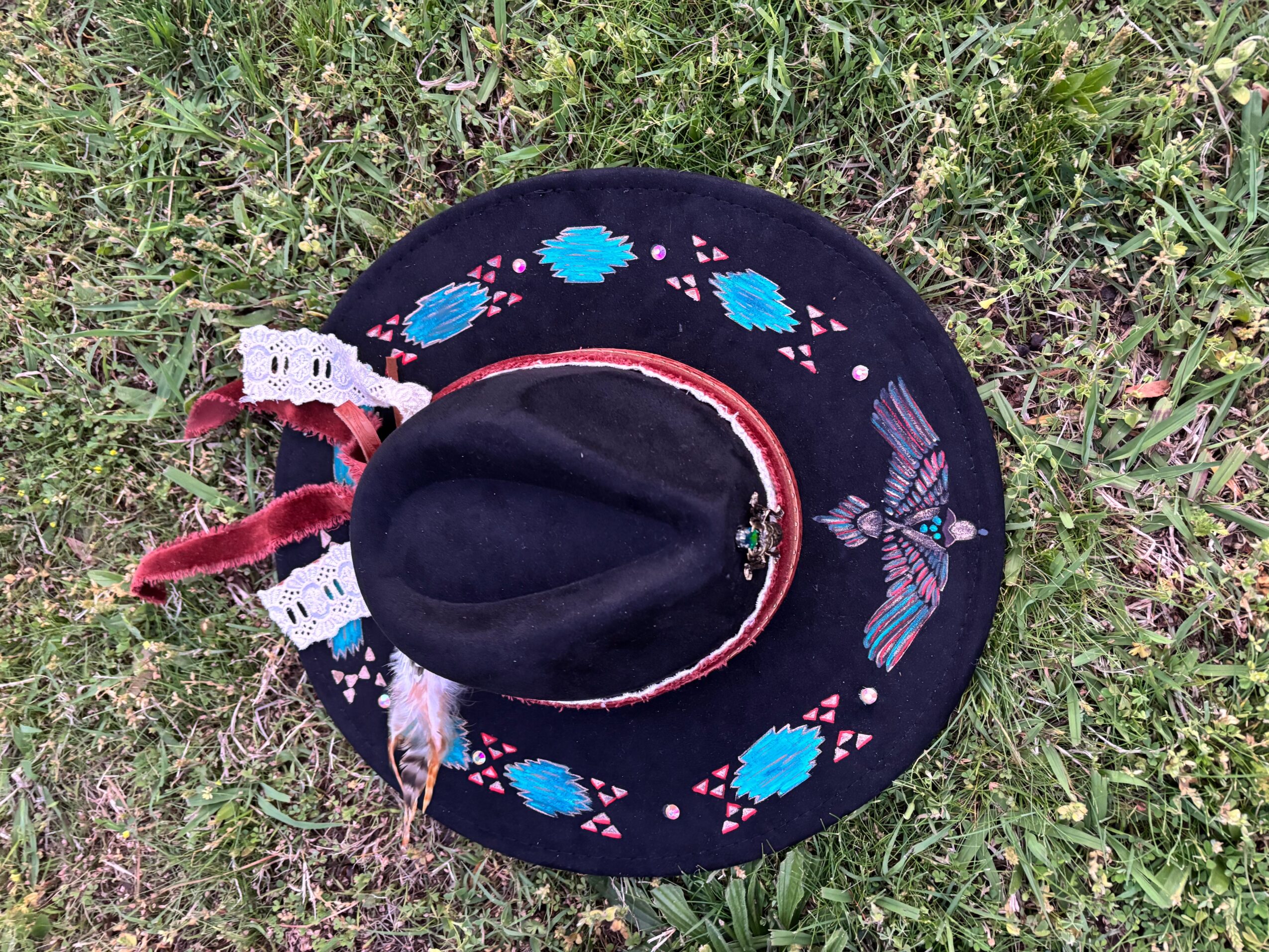 A black decorated hat with turquoise and pink patterns lies on grass.