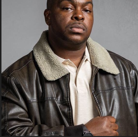 Man wearing a leather jacket with a shearling collar, white shirt, and serious expression, against a gray background.