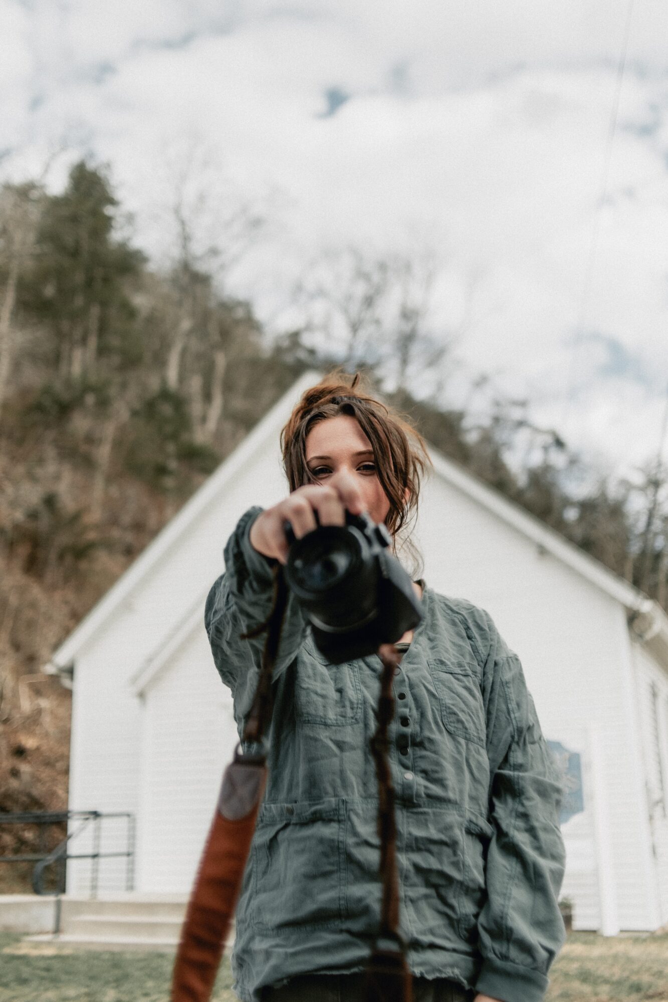 Person holding camera, standing outdoors in front of a white building with trees and cloudy sky, face partially visible.