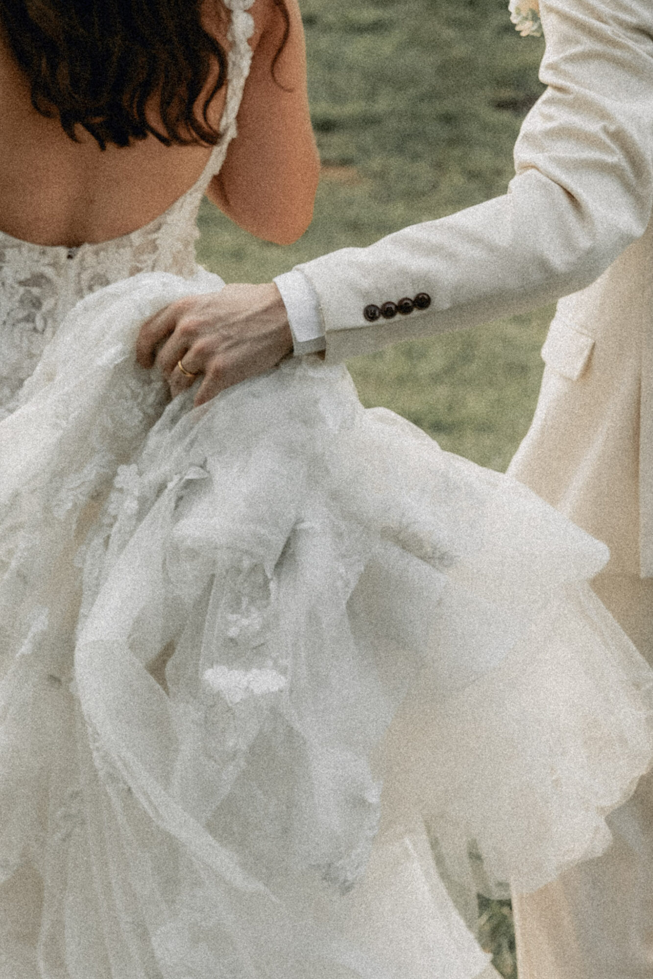 A person in a white suit holds a woman's wedding dress outdoors, with grass in the background.
