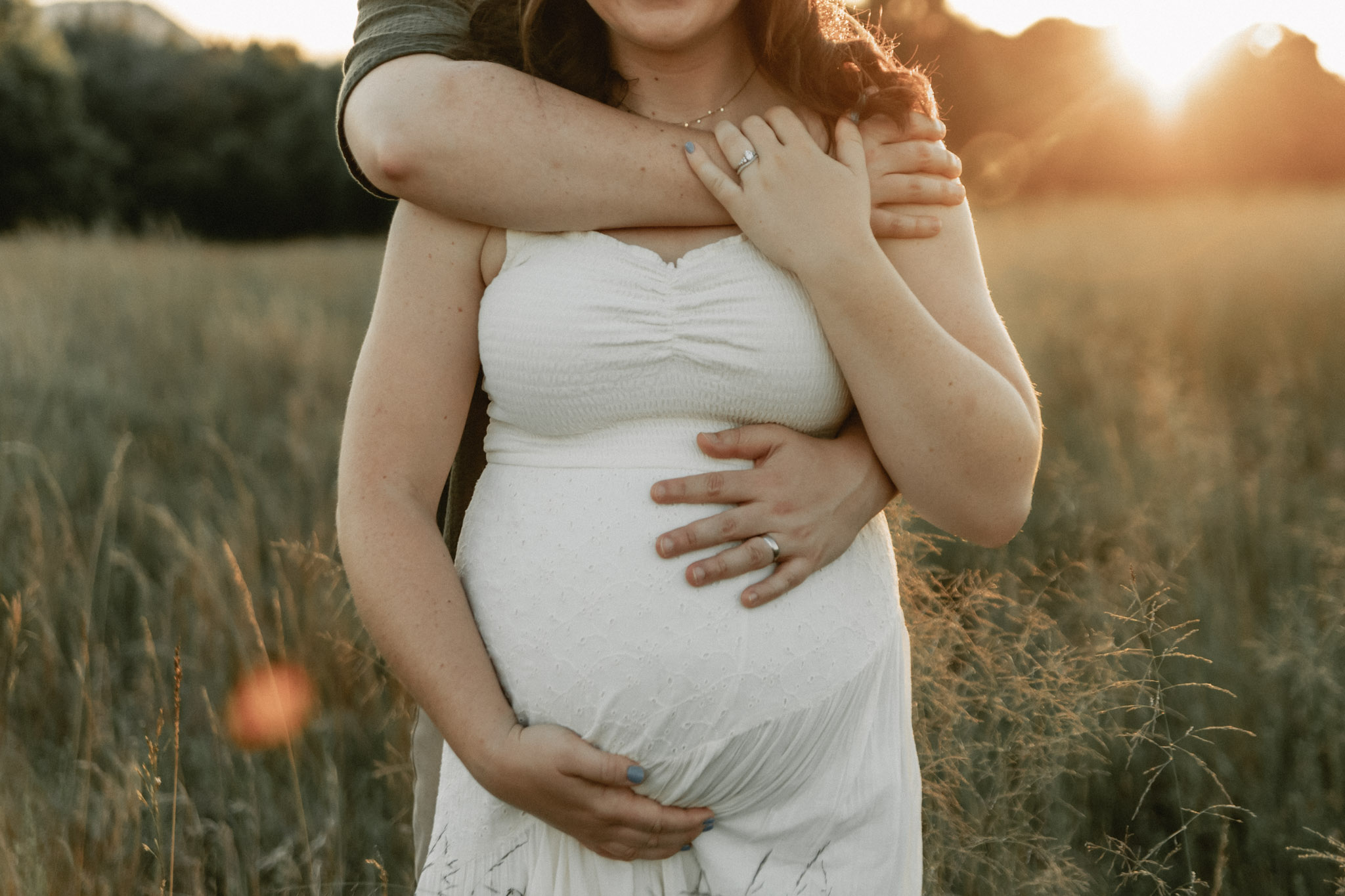 Pregnant woman in a white dress with a man embracing her outdoors at sunset, holding her belly.